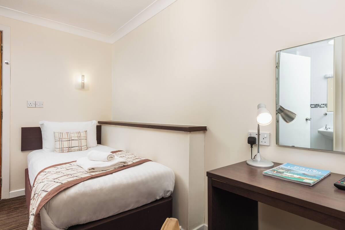 A hotel room with a neatly made bed, a desk with a lamp, a book, and a mirror.