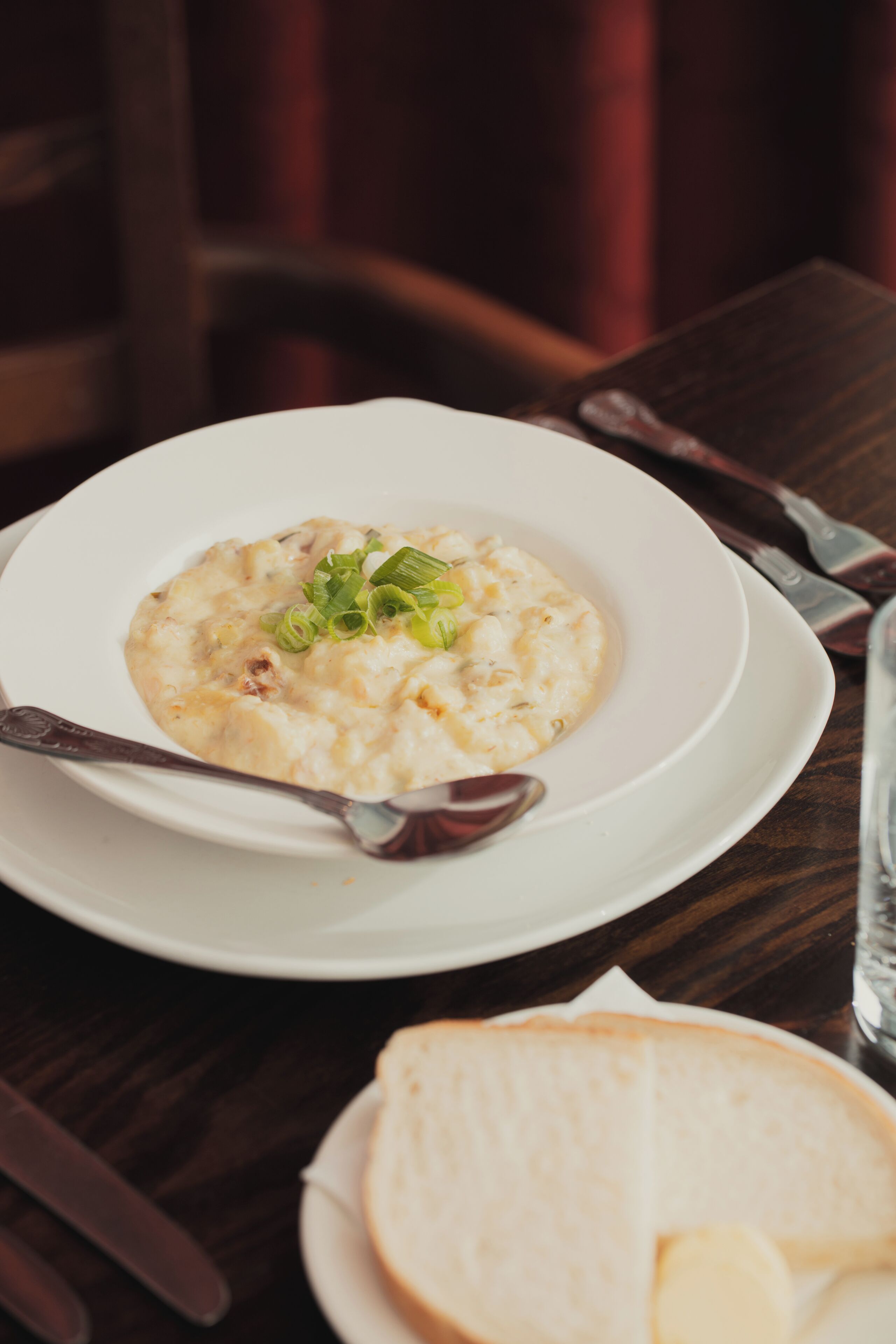 A white bowl of creamy soup with green garnish on a white plate, accompanied by a slice of bread and a spoon on a wooden table.