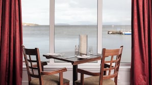 A dining table with two chairs, a menu, and a glass of water, positioned by a large window overlooking a body of water and a distant island. - Dunollie Hotel (Strath)