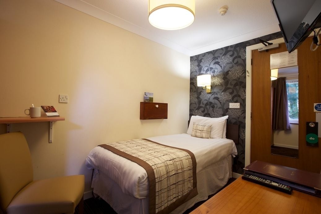 A hotel room with a bed, bedside table, chair, and a television mounted on the wall.