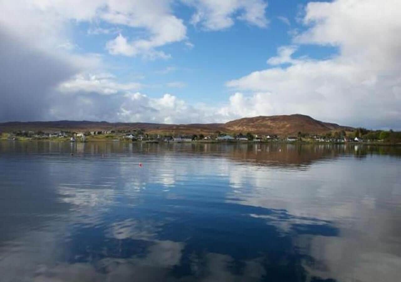 Un lago tranquilo con una montaña al fondo y una hilera de edificios a lo largo de la orilla.