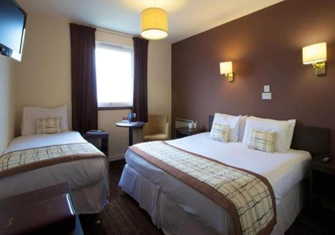 A hotel room with two beds, a small table, a chair, and a window with curtains.