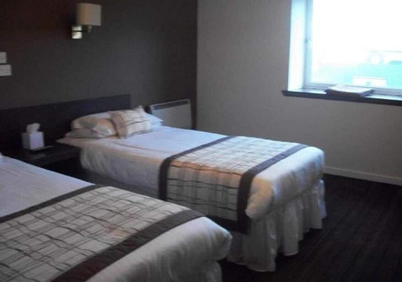 A hotel room with two single beds, a nightstand, a lamp, and a window with a view.