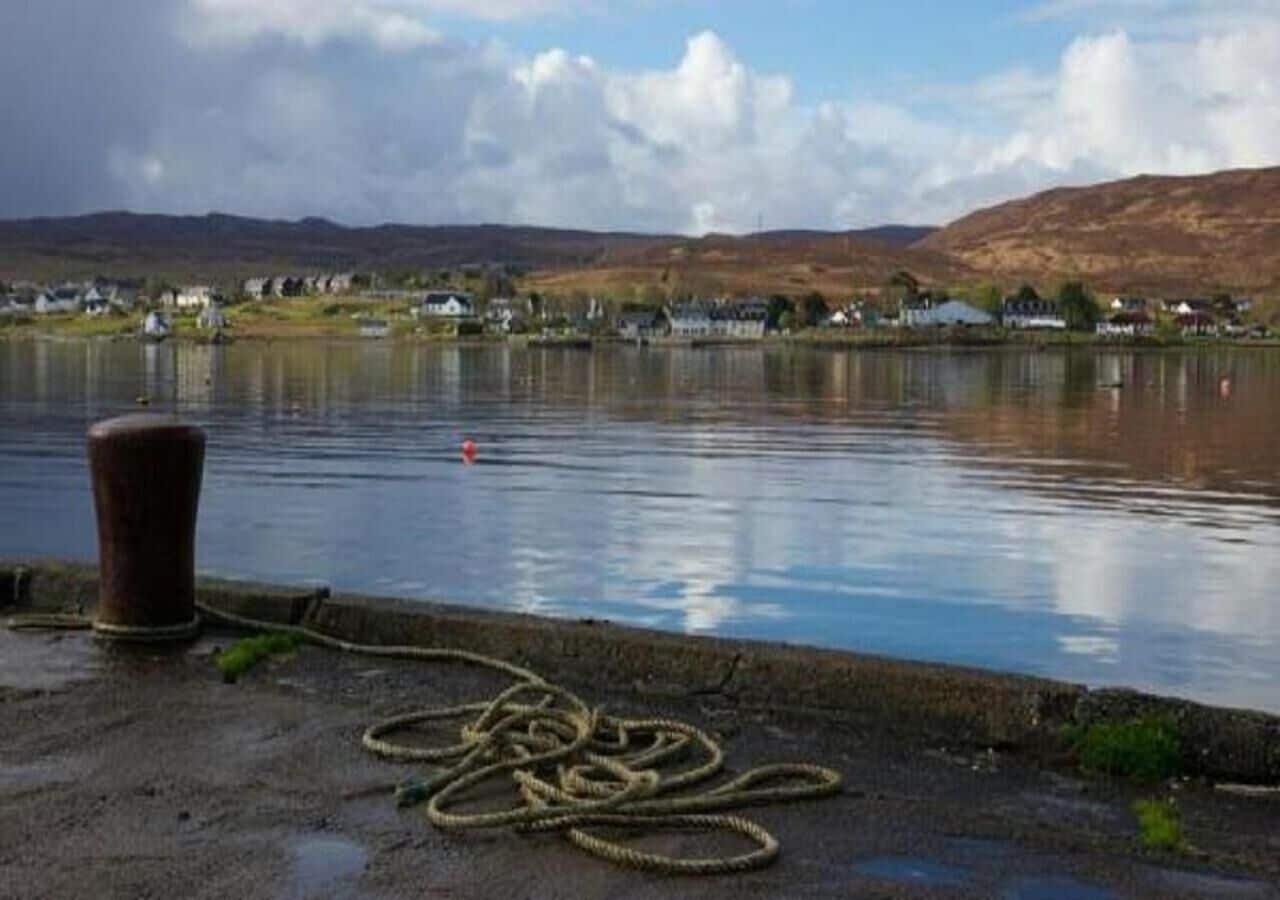 Ein ruhiger See mit einer Seilrolle und einem Anker am Ufer, umgeben von Häusern und Hügeln im Hintergrund.