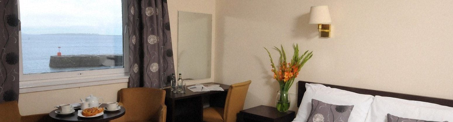 Een hotelkamer met uitzicht op zee, een eettafel met theeservies, een vaas met bloemen en een bedlamp.