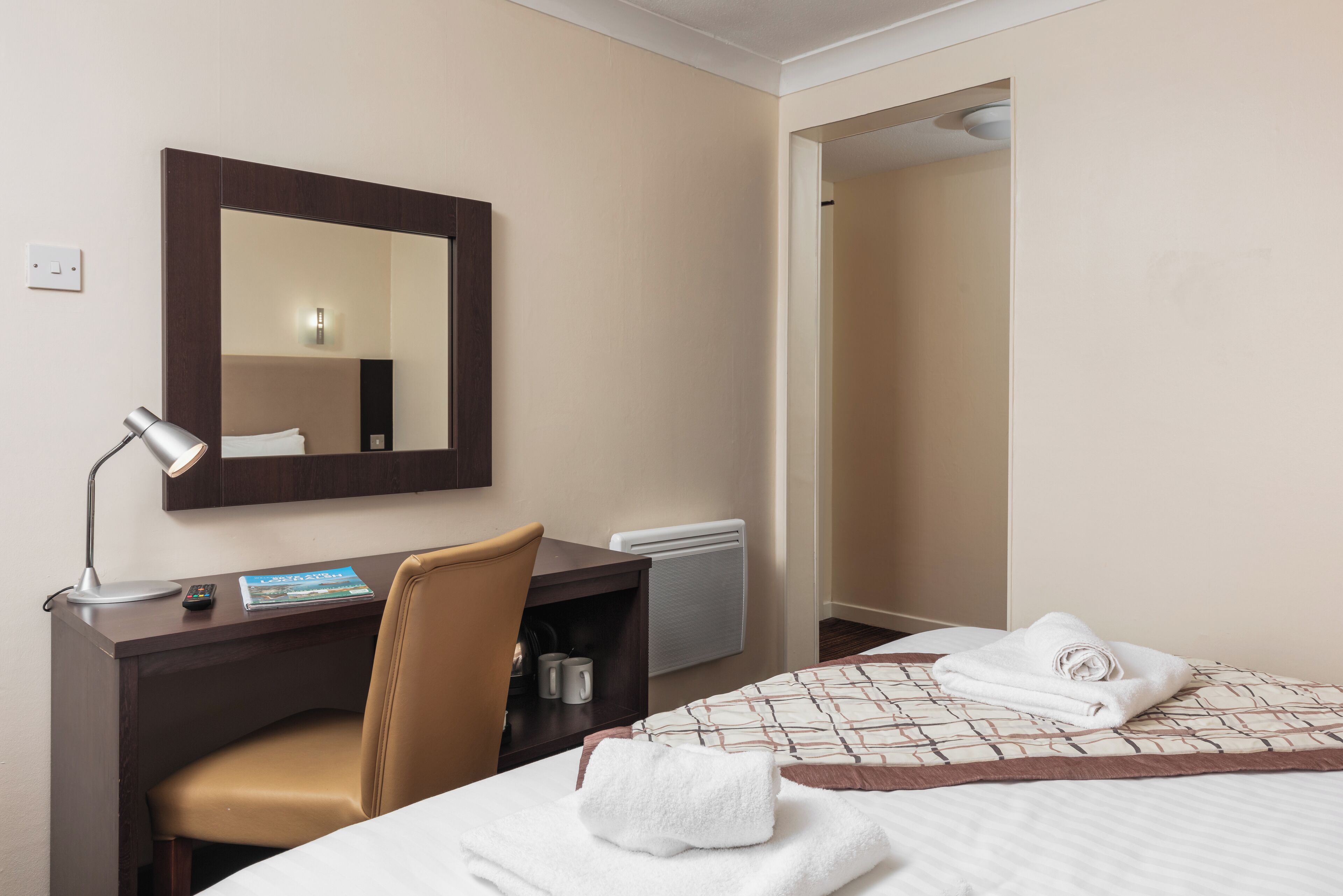 A hotel room with a bed, a desk with a chair, a mirror, and a radiator.