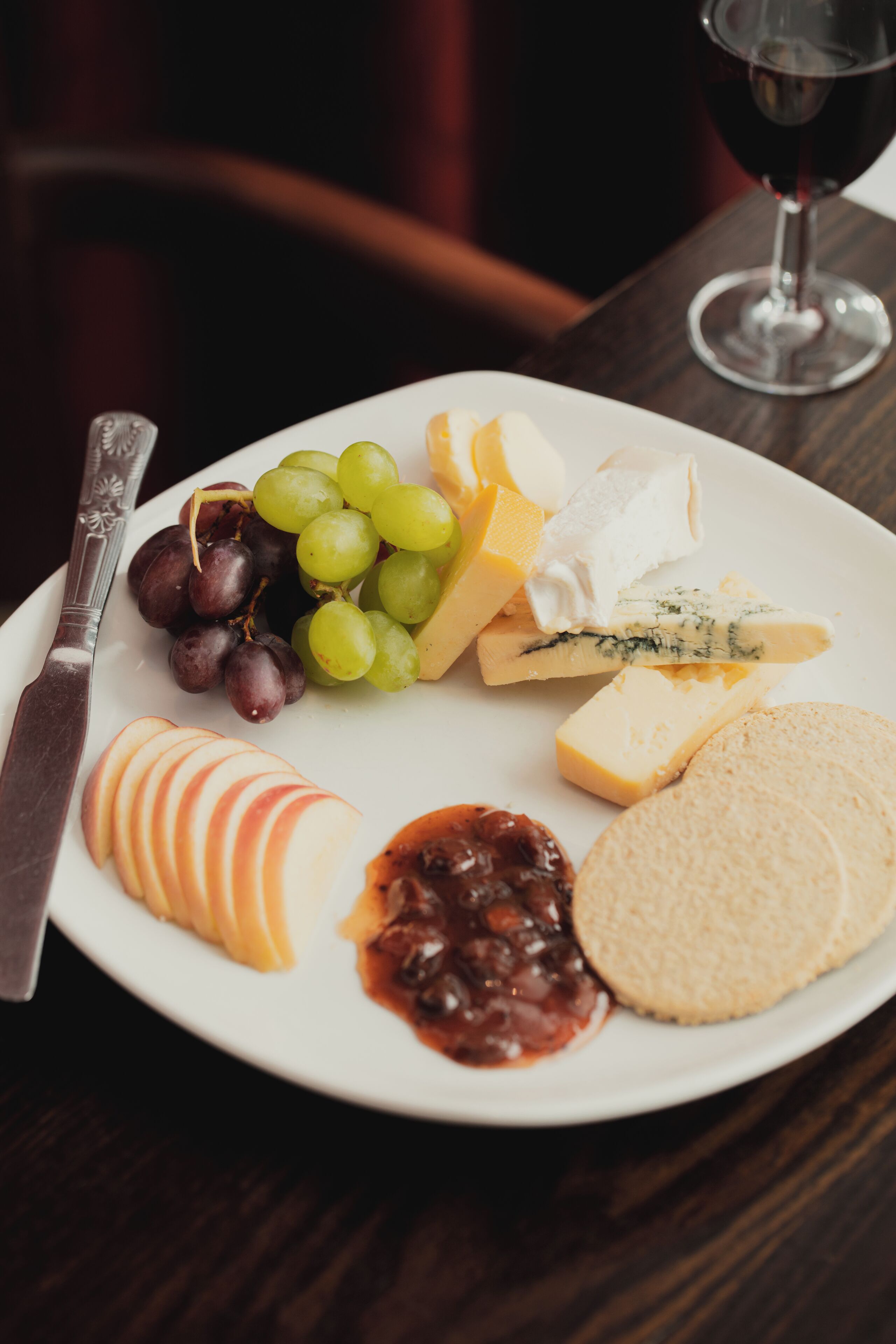 Una tabla con varios quesos, uvas, rodajas de manzana y galletas sobre una mesa.