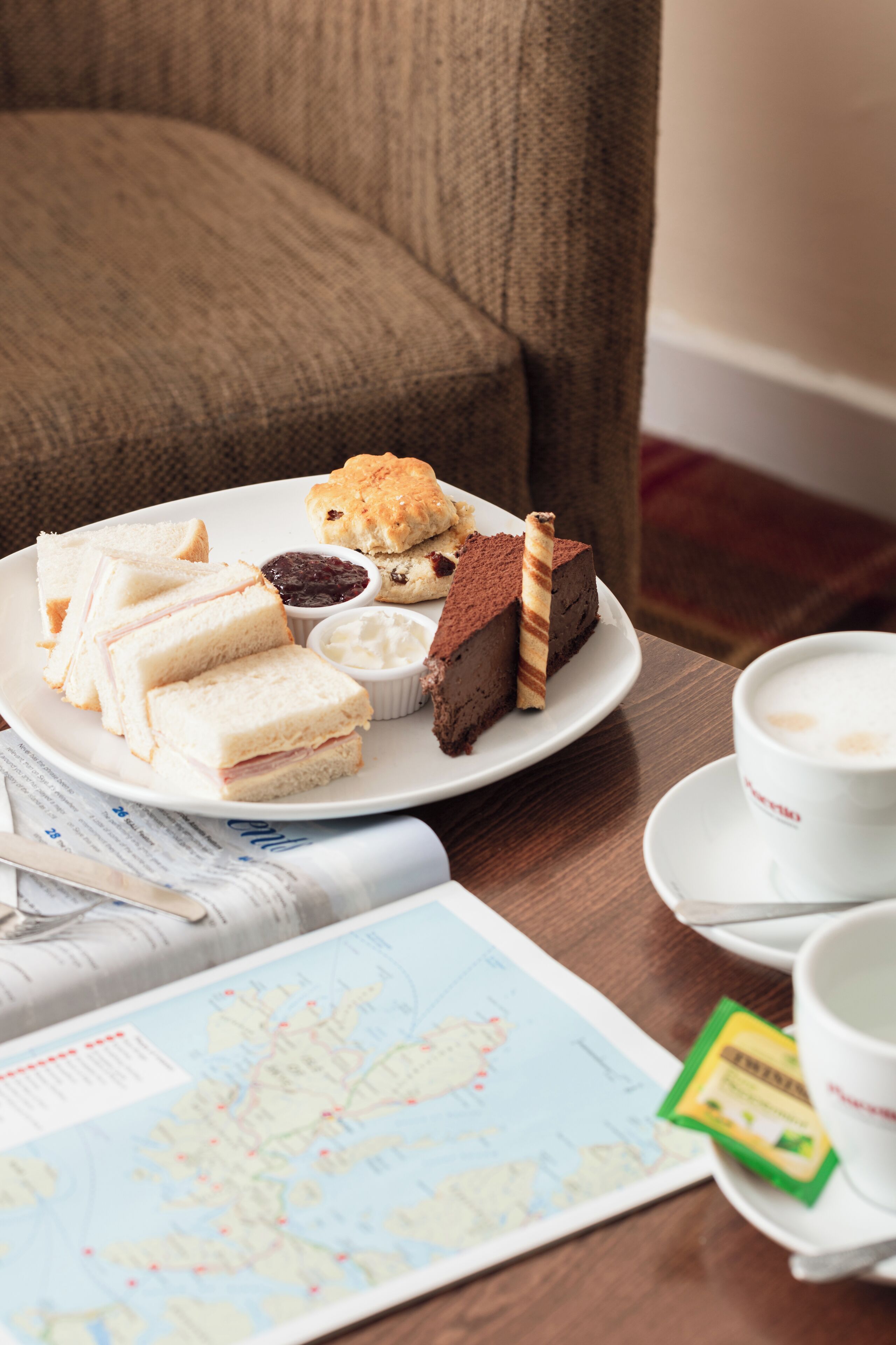A plate of sandwiches, scones, and a slice of cake on a table with a map and a cup of coffee.