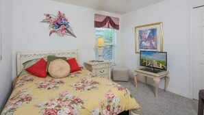 A bedroom with a bed, a ceiling fan, a window with curtains, a bedside table with a lamp, a television on a stand, and a framed picture on the wall.