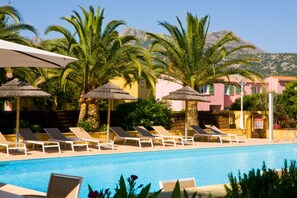 Un área de piscina con sillas de descanso y sombrillas, rodeada de palmeras y edificios con fachadas rosadas y amarillas.
