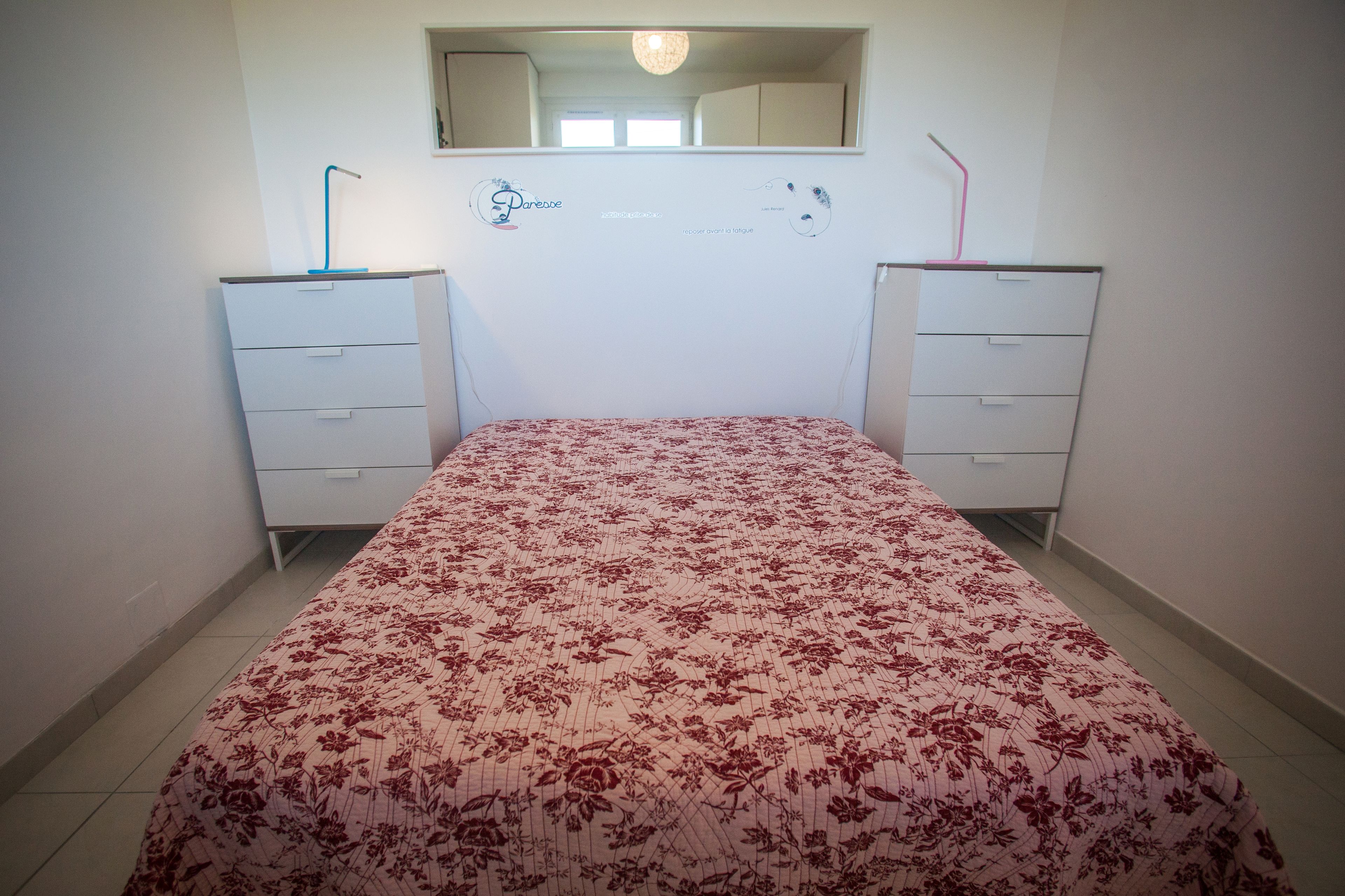 A bedroom with a floral bedspread, two white dressers, a mirror above the bed, and a wall decal.