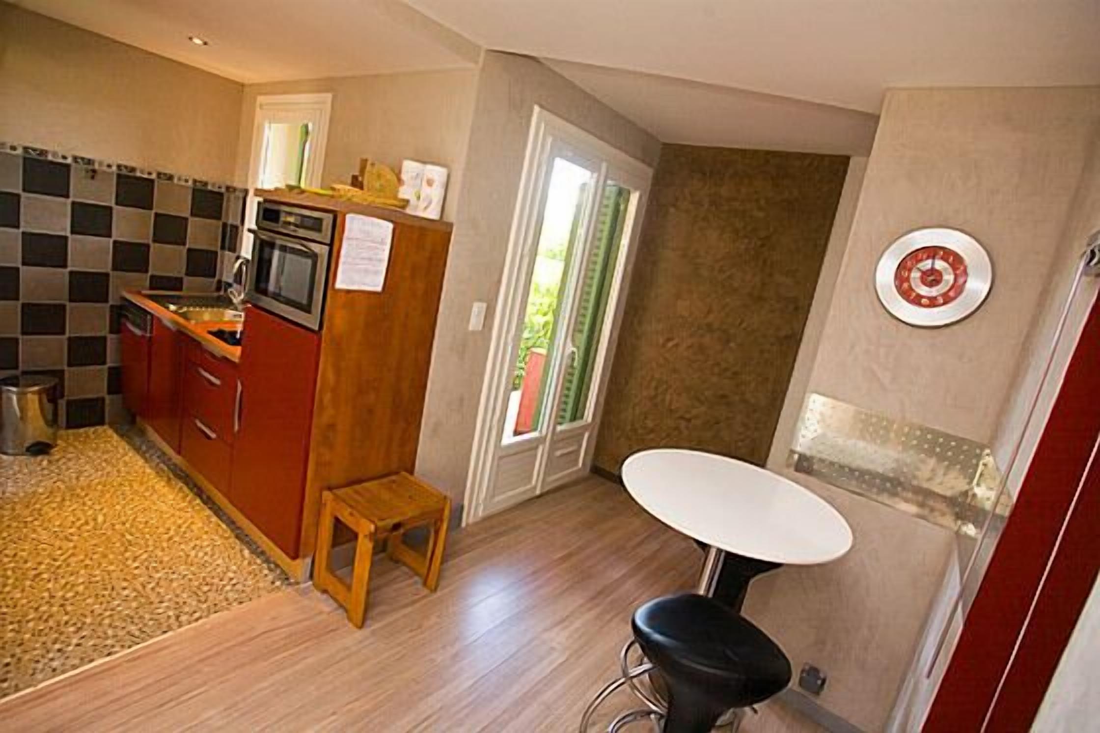 A compact kitchen with a stove, oven, and wooden cabinets. There's a small stool and a white table with a black chair.