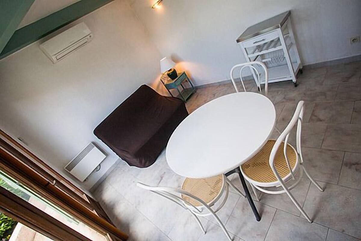 Una habitación pequeña y bien iluminada con una mesa y sillas blancas, un sofá marrón oscuro, un mueble blanco pequeño y una ventana con persianas.