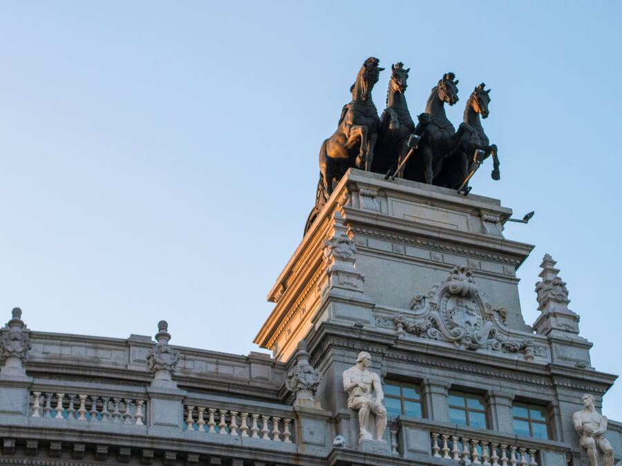 Un edificio con una llamativa escultura de cuatro caballos en la parte superior, detalles arquitectónicos elaborados y un asta de banderas.