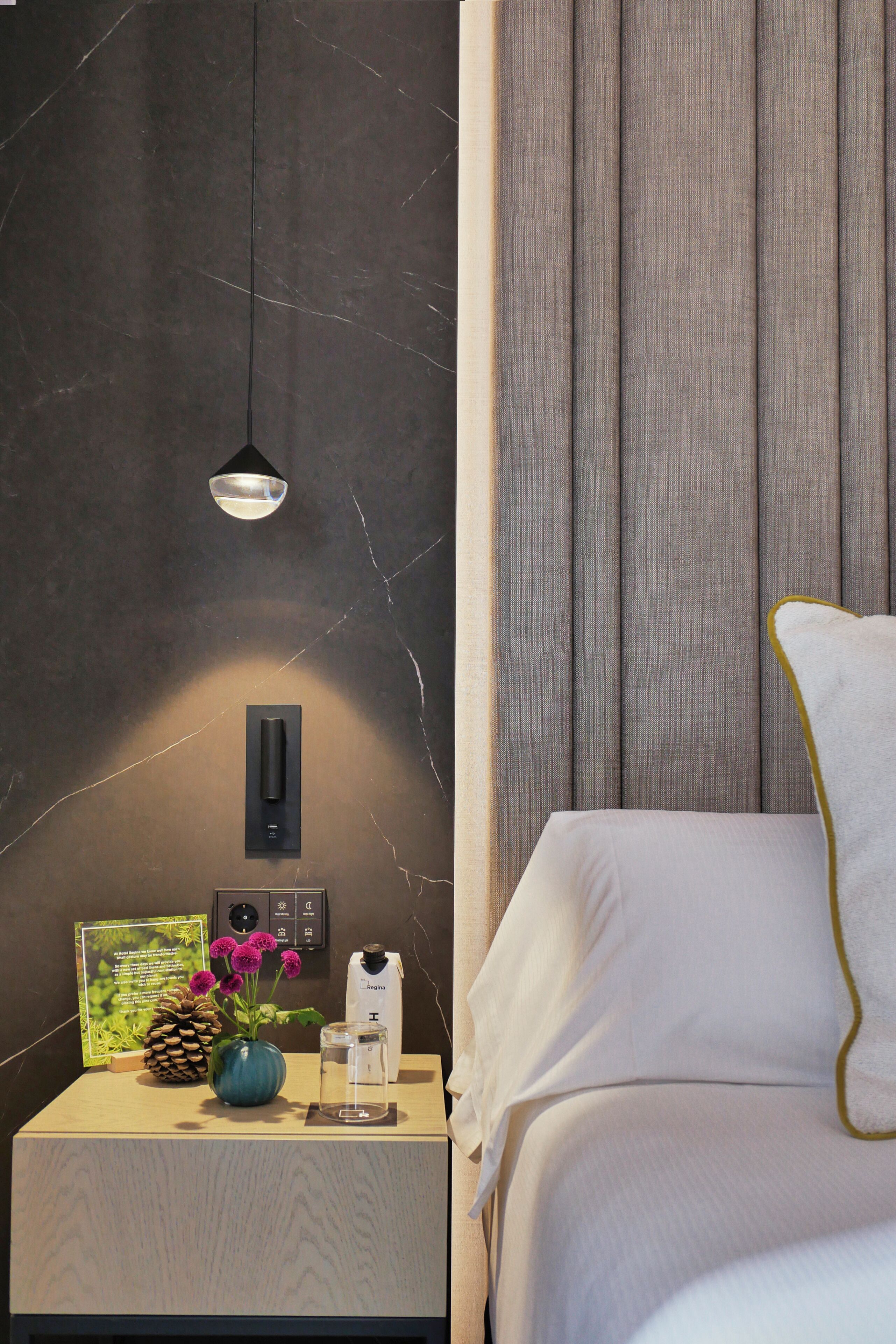 Una camera d'albergo con un letto, un comodino, un vaso con fiori, un quadro incorniciato, una bottiglia e un lampadario a parete.