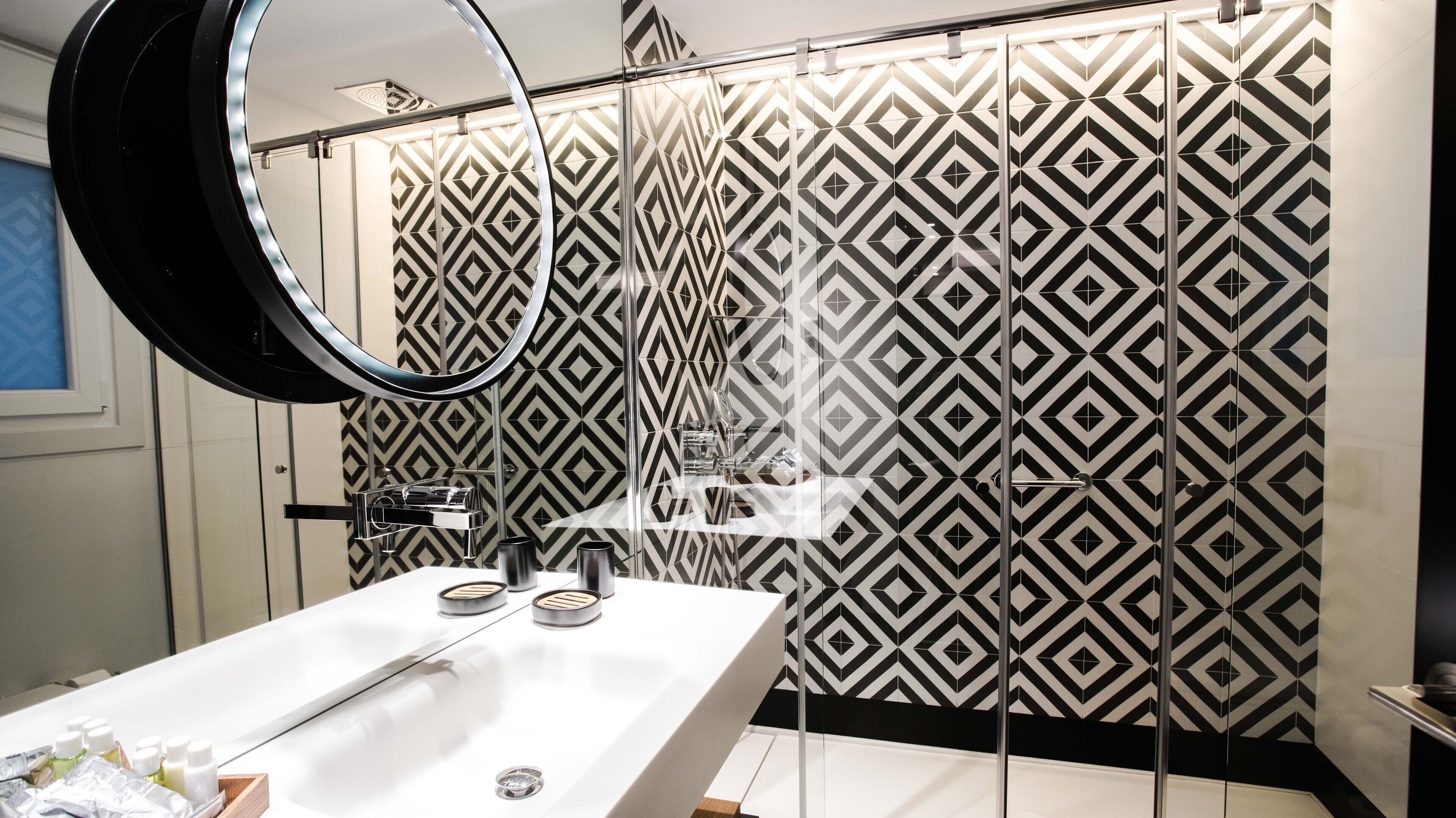 Un baño moderno con un revestimiento mural de diseño geométrico, un lavamanos blanco con espejo y una zona de ducha.