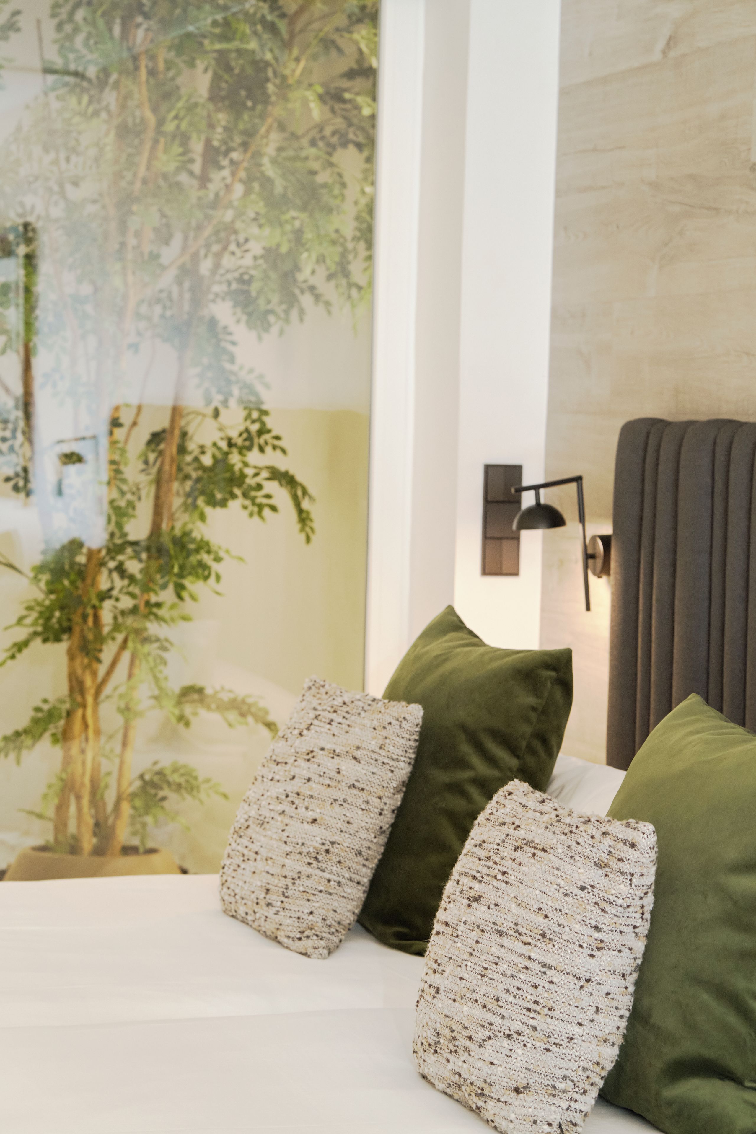 Un dormitorio con cama, almohadas, una lámpara fijada a la pared y un ventanal con vistas a árboles.