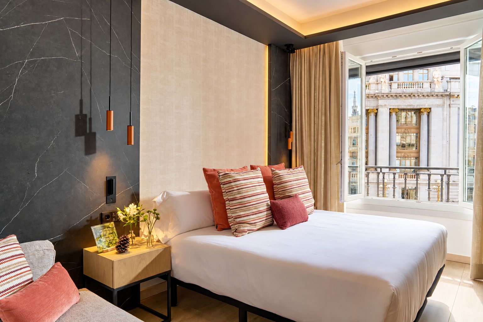 Camera d'albergo con un letto grande, un comodino, un vaso con fiori e vista su un edificio dalla finestra.