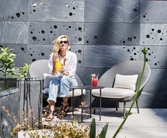 A person sitting on a grey chair with a drink on a small table, in front of a modern wall with circular cutouts.