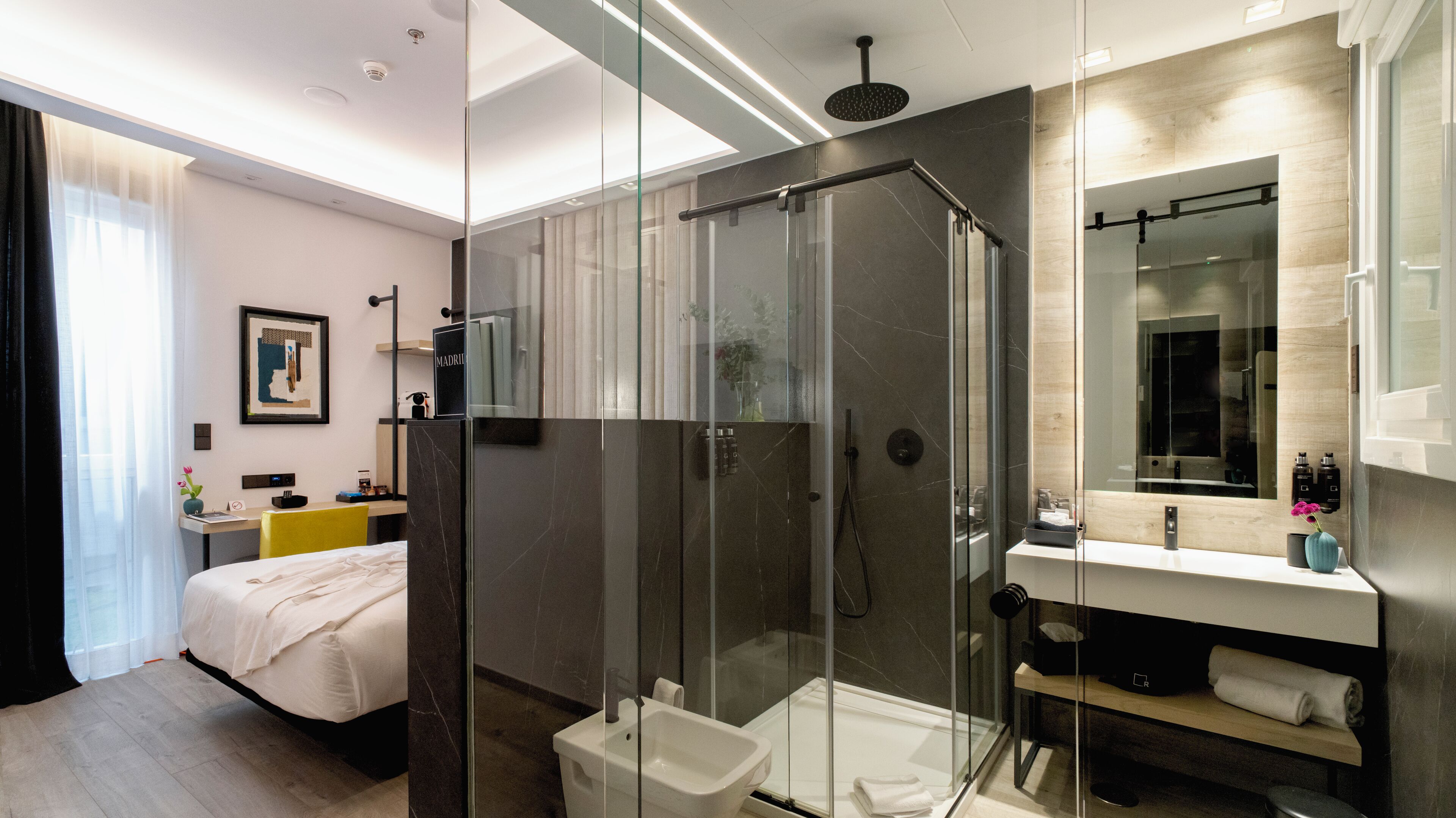 Une chambre d’hôtel moderne dotée d’une douche en verre, d’un lit avec du linge de lit blanc, d’une table de chevet avec une lampe et d’un grand miroir au-dessus du lavabo.