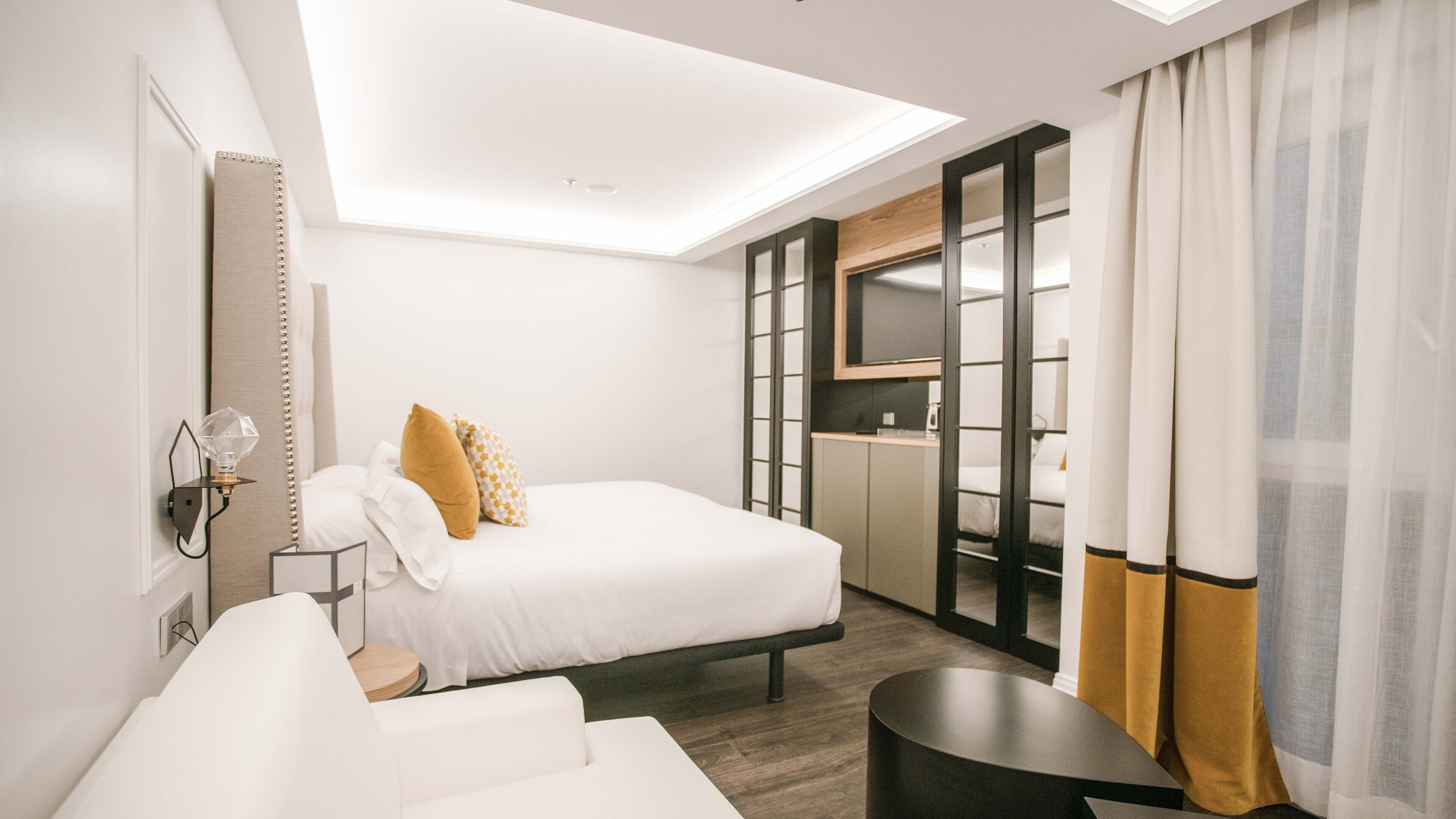 A modern hotel room with a bed, a desk, a chair, a small table, and a kitchenette area.