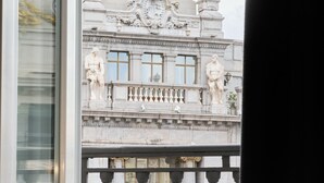 Un balcón con alimentos para el desayuno, dos vasos de jugo y vistas a una estatua y la fachada de un edificio.