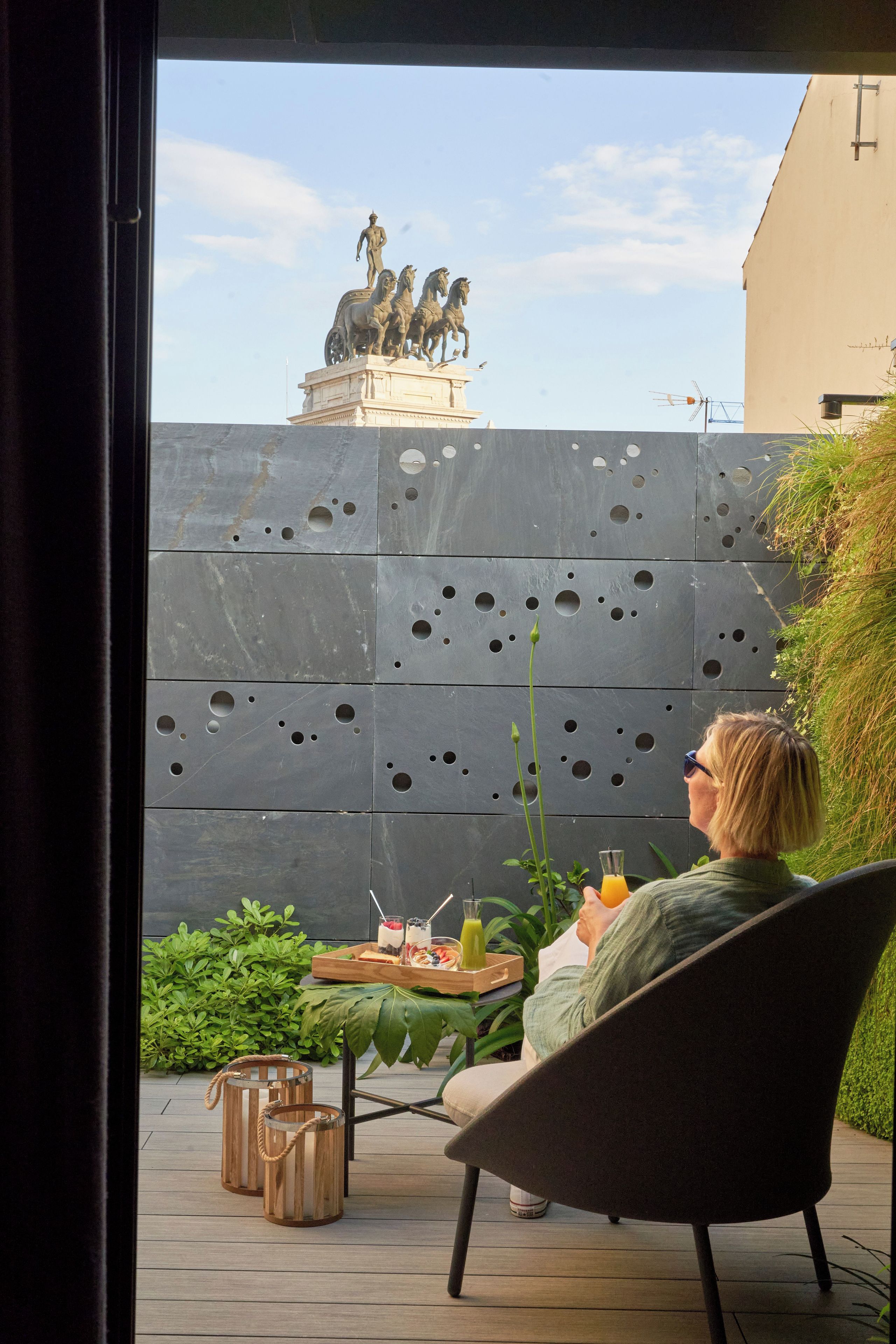 Una persona sentada en una mesa al aire libre con una bandeja de comida y bebidas, con vista a un patio que tiene una estatua y una pared moderna con círculos.