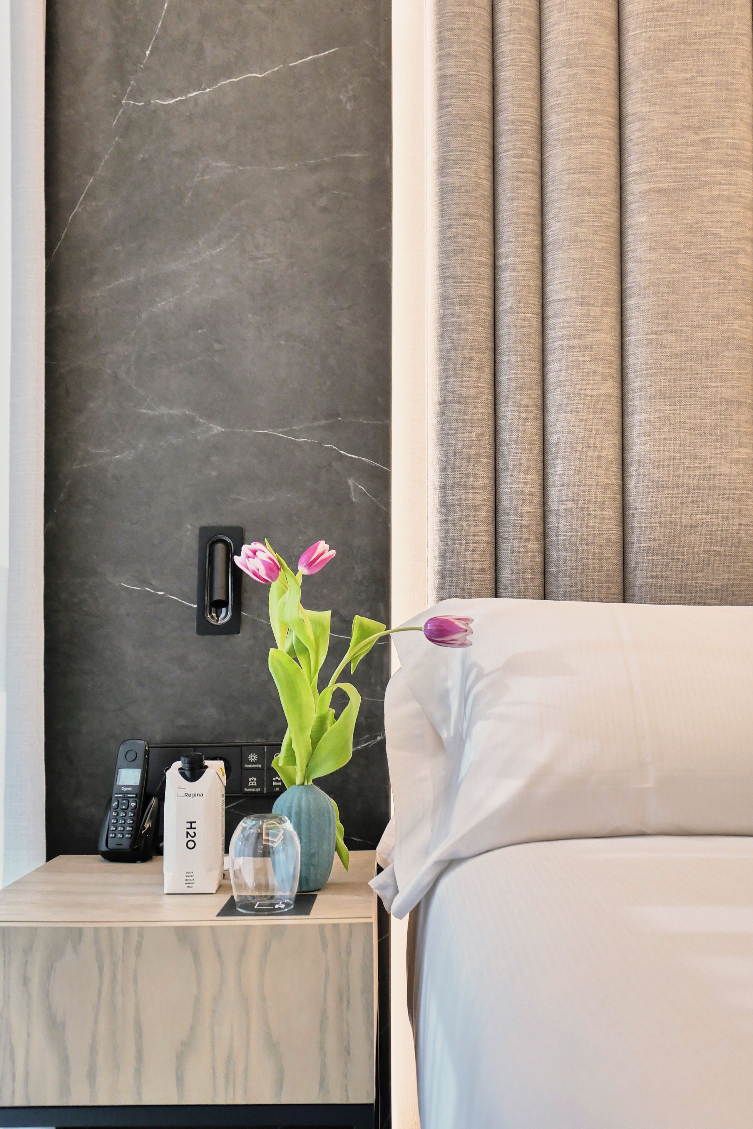 Ein Hotelzimmer mit Marmorwand, einem Nachttisch mit Telefon, einer Wasserflasche und einer Vase mit Blumen.