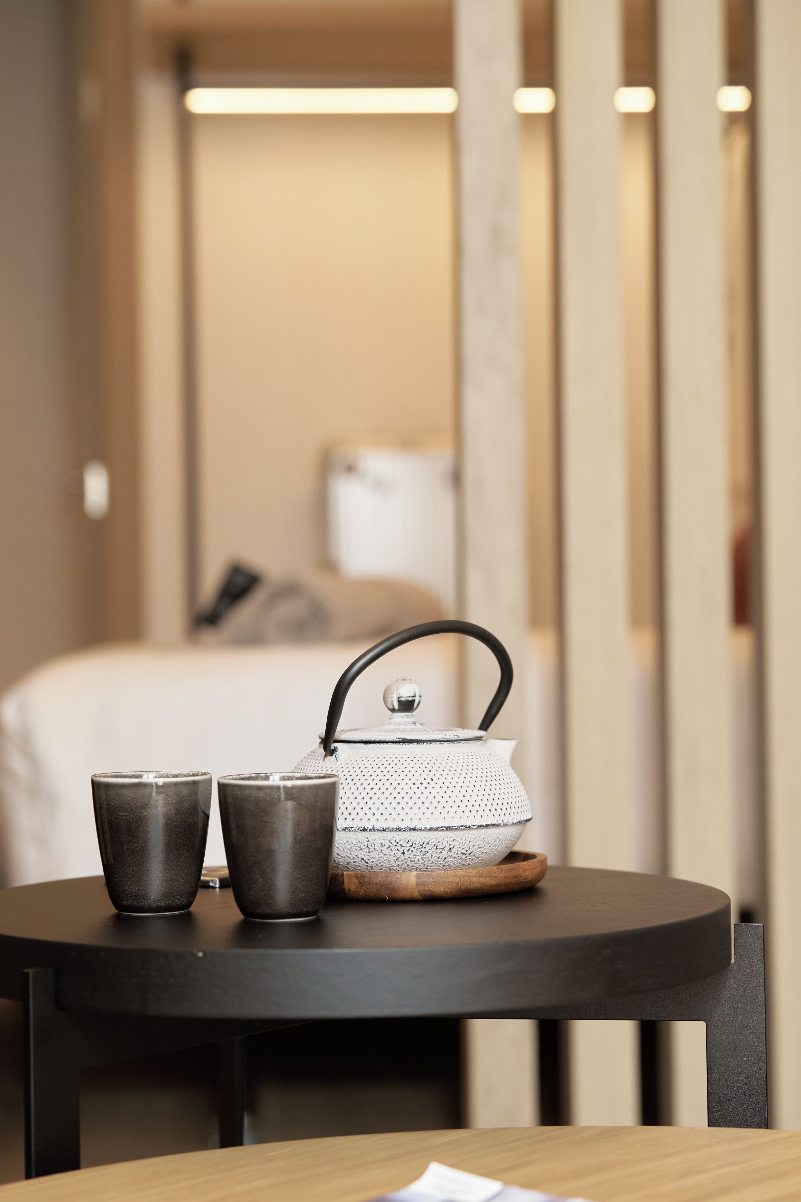 Una mesa redonda con una tetera y dos tazas, una cama y una mesita de noche al fondo.