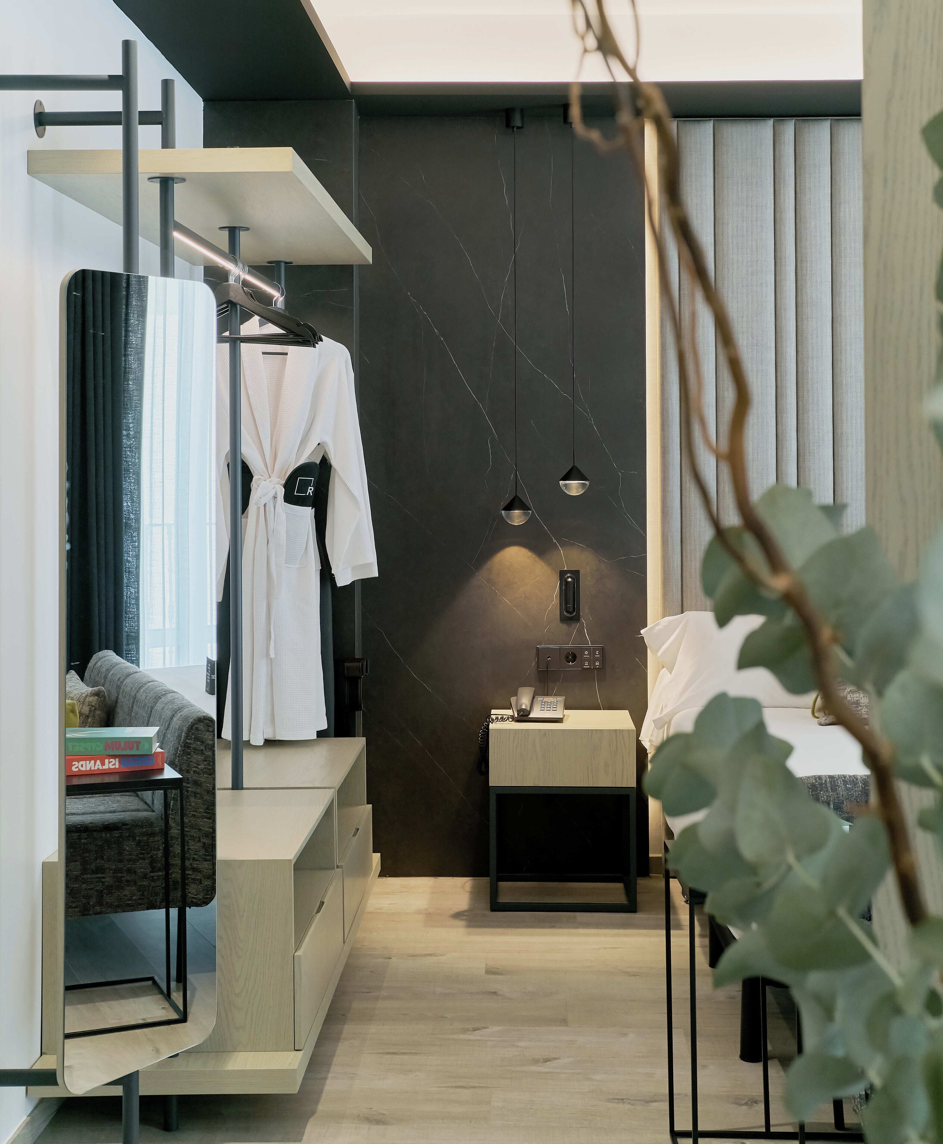 A modern, compact hotel room with a minimalist design, featuring a bed, bedside table, and a seating area with a sofa and books.