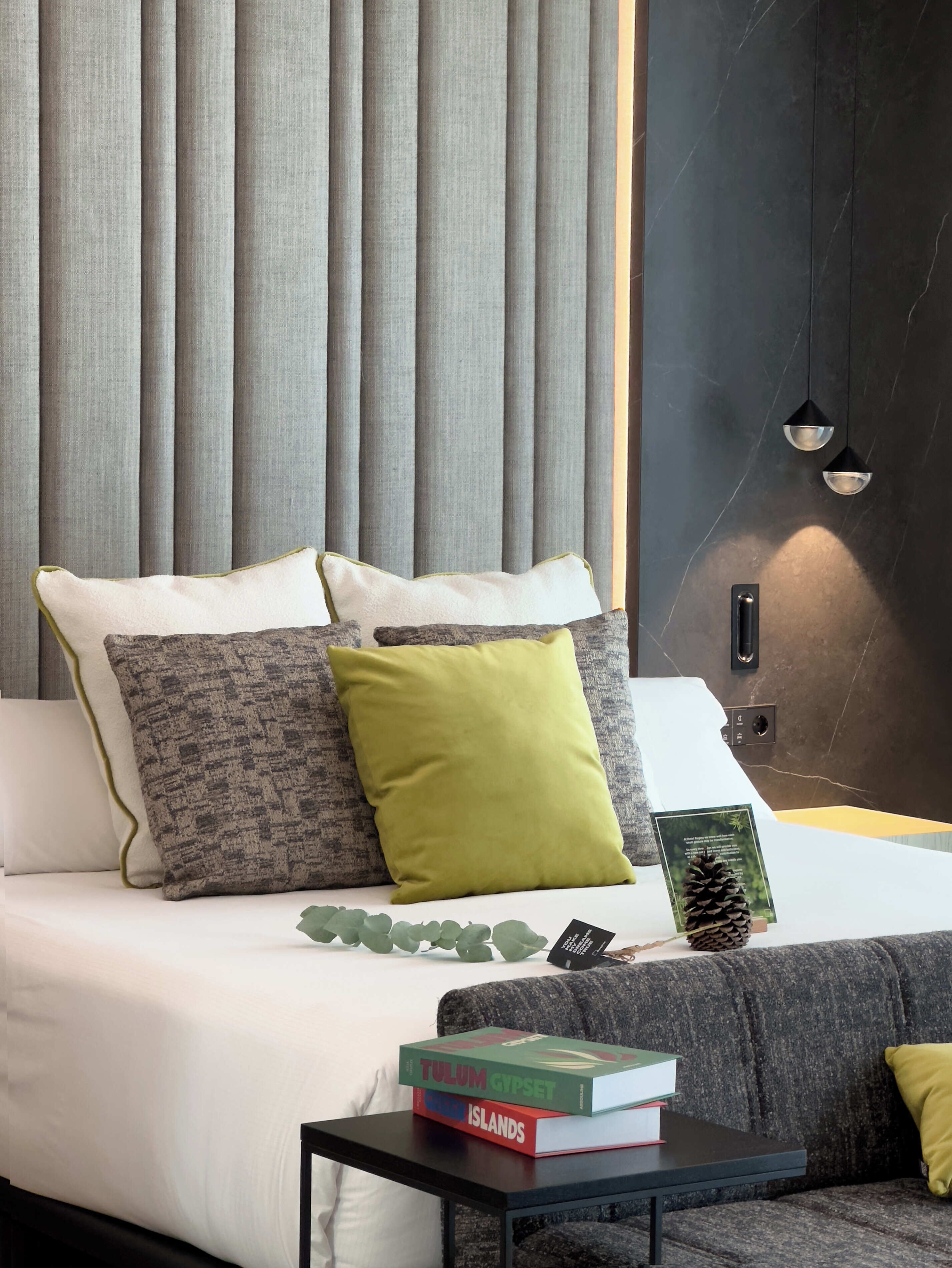 Un dormitorio moderno con cama, almohadas, una mesita de noche con libros y una lámpara fijada a la pared.