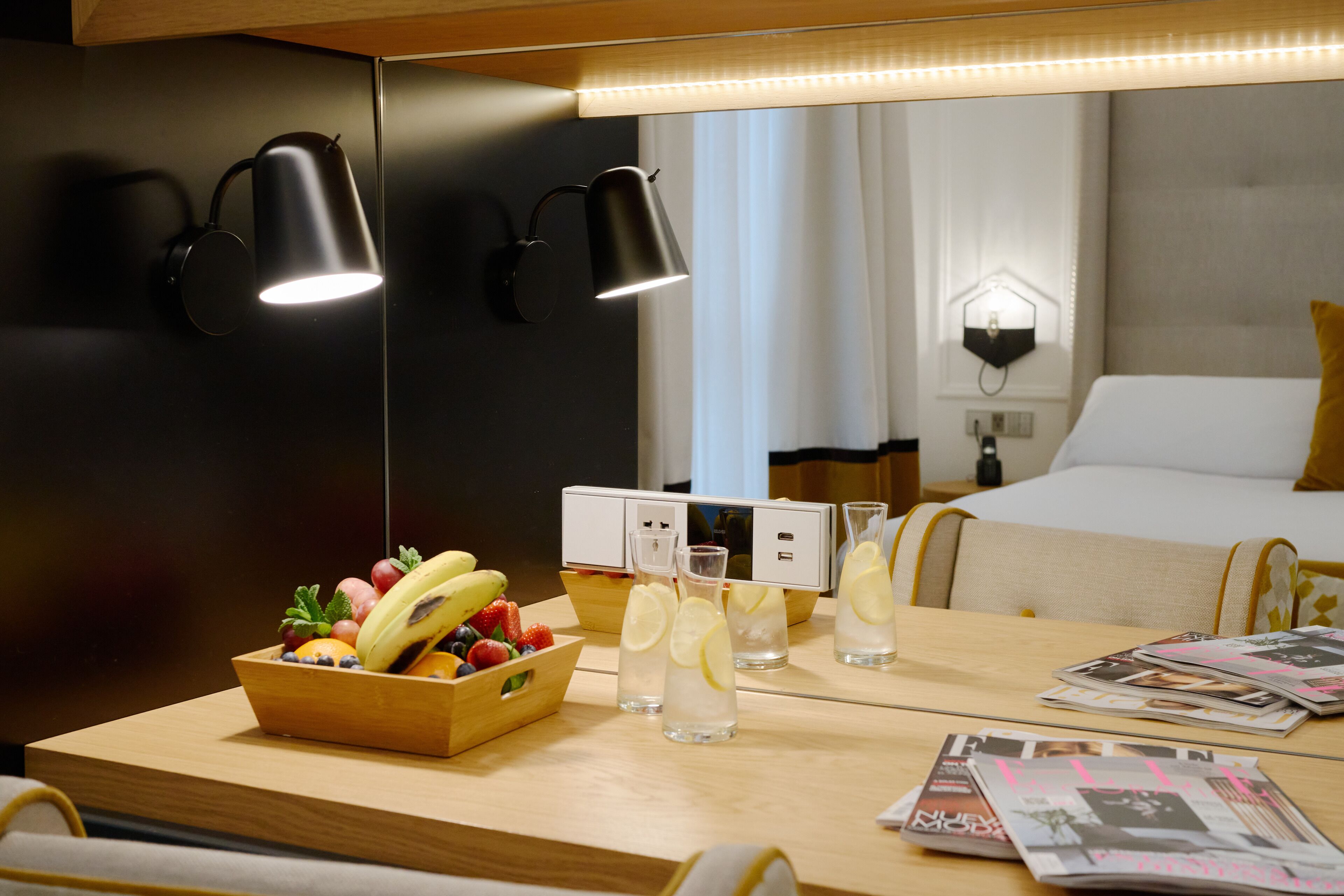 Habitación de hotel con una mesa de madera, frutero, vasos de agua y revistas.