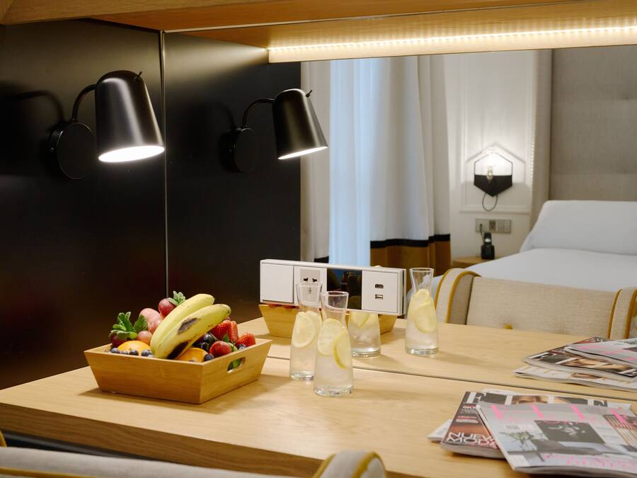 Habitación de hotel con una mesa de madera, frutero, vasos de agua y revistas.