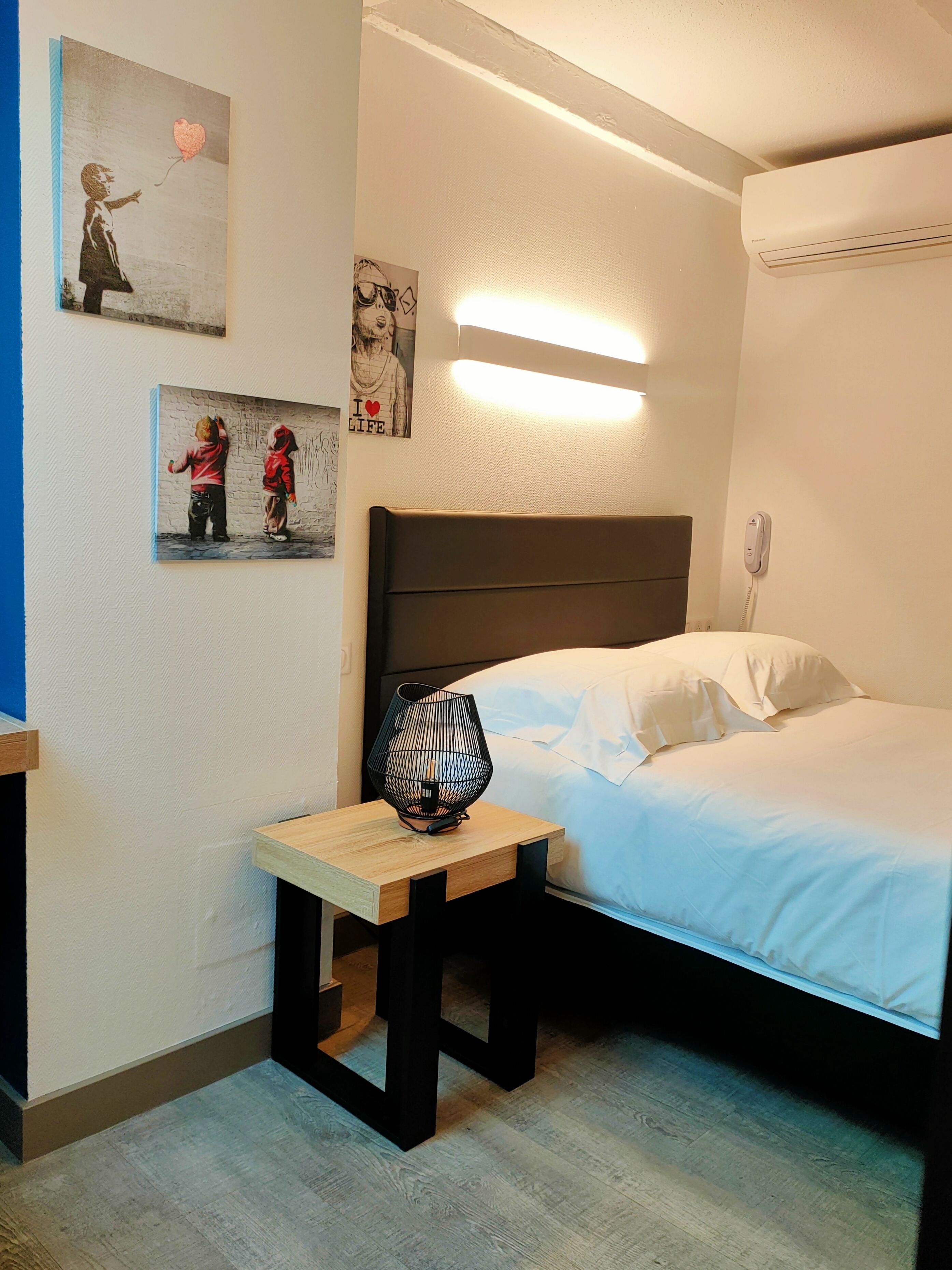 A bedroom with a bed, bedside table, lamp, wall art, and an air conditioning unit.