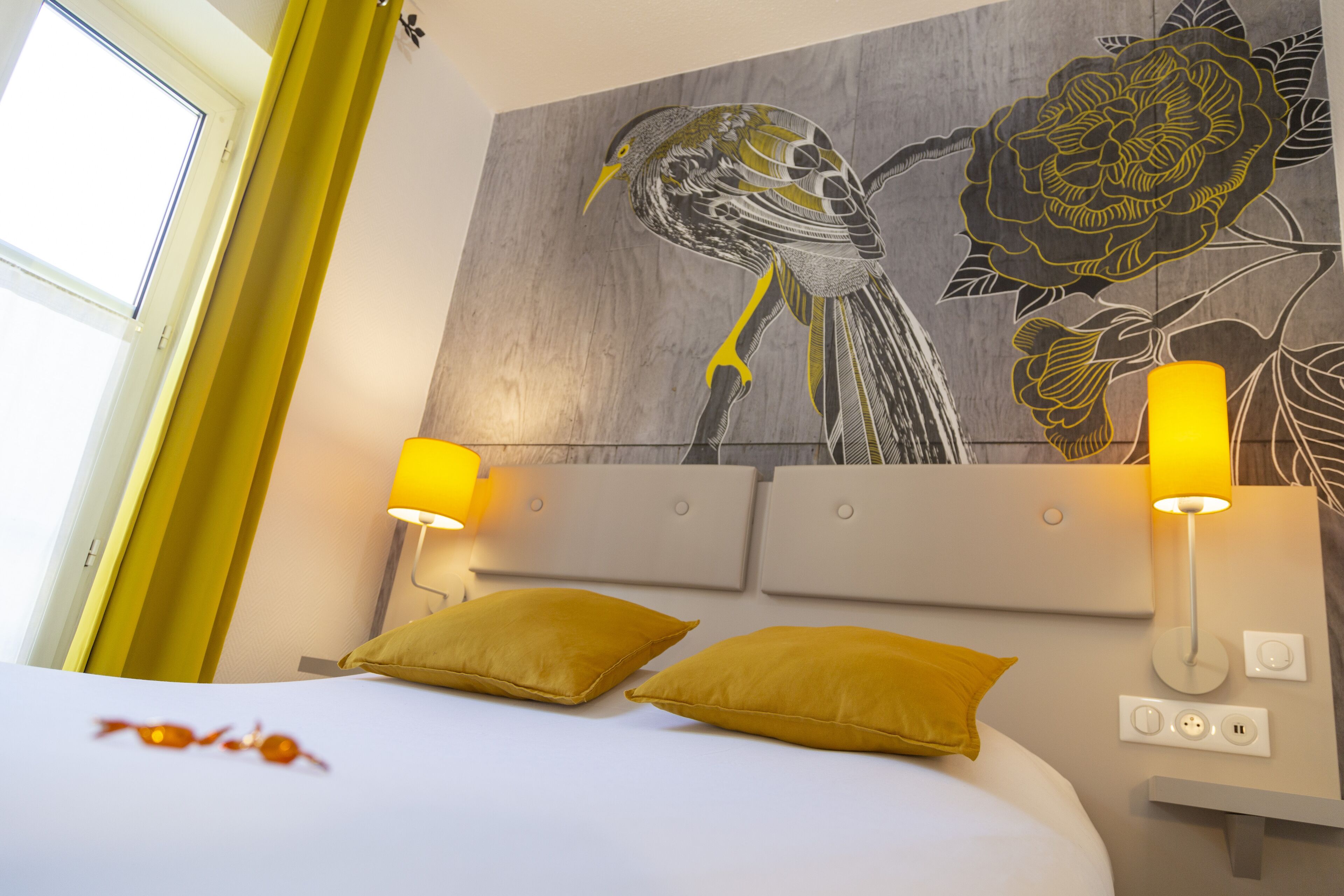 A hotel room with a bed, two yellow pillows, a wall-mounted lamp, and a painting of birds and flowers.