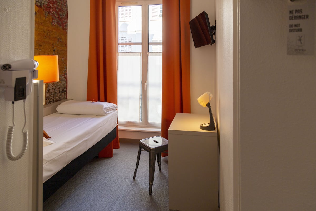 A hotel room with a bed, a desk, a chair, a lamp, a telephone, and a window with curtains.