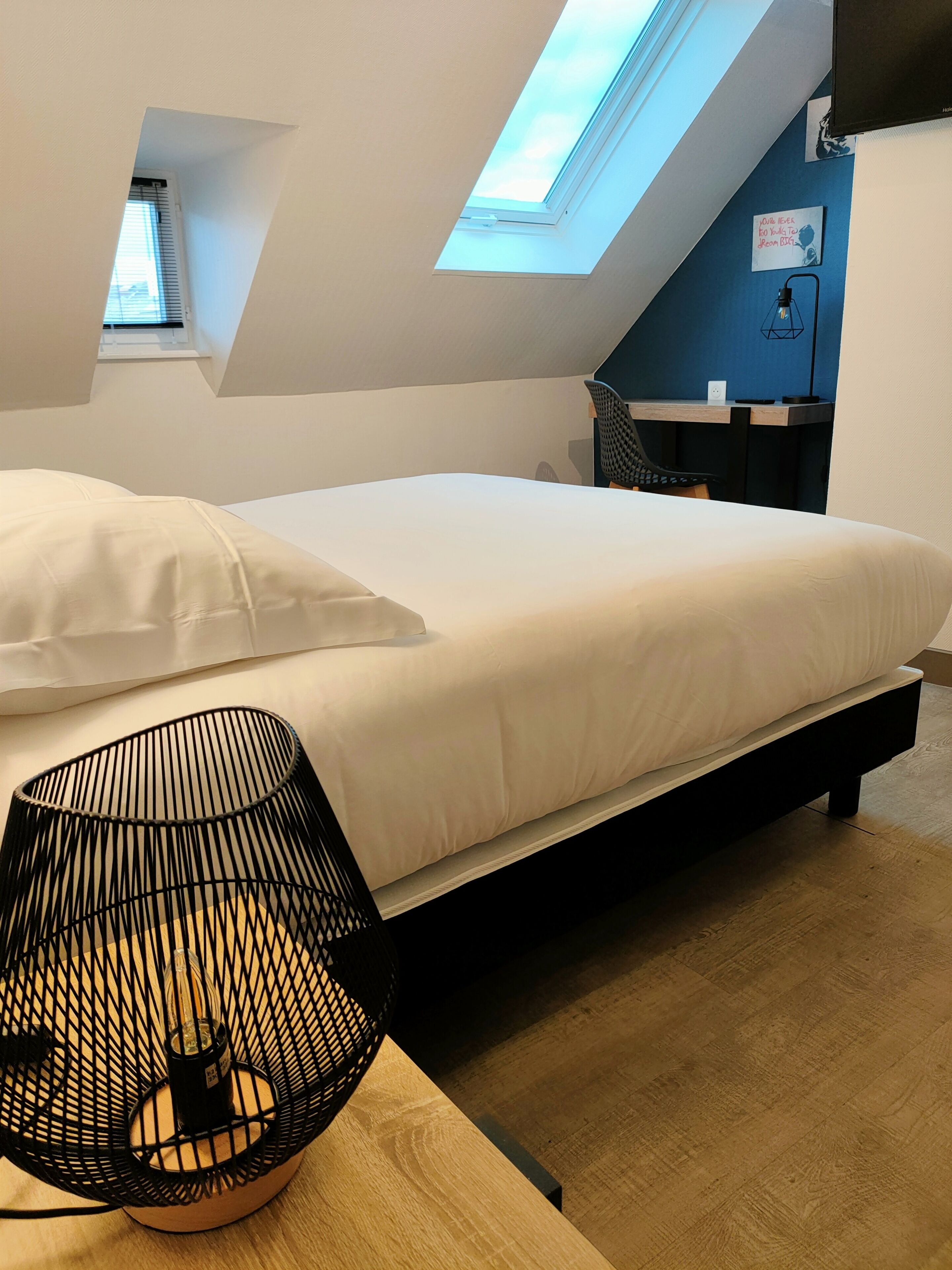 Une chambre moderne avec une lucarne, un lit recouvert de linge de lit blanc, une lampe en métal noir et un petit bureau avec une chaise.