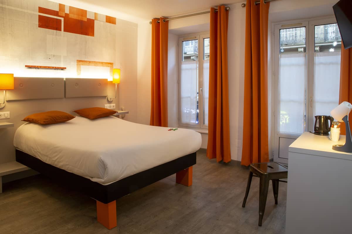 A modern hotel room with a large bed, a desk with a lamp, and orange curtains.