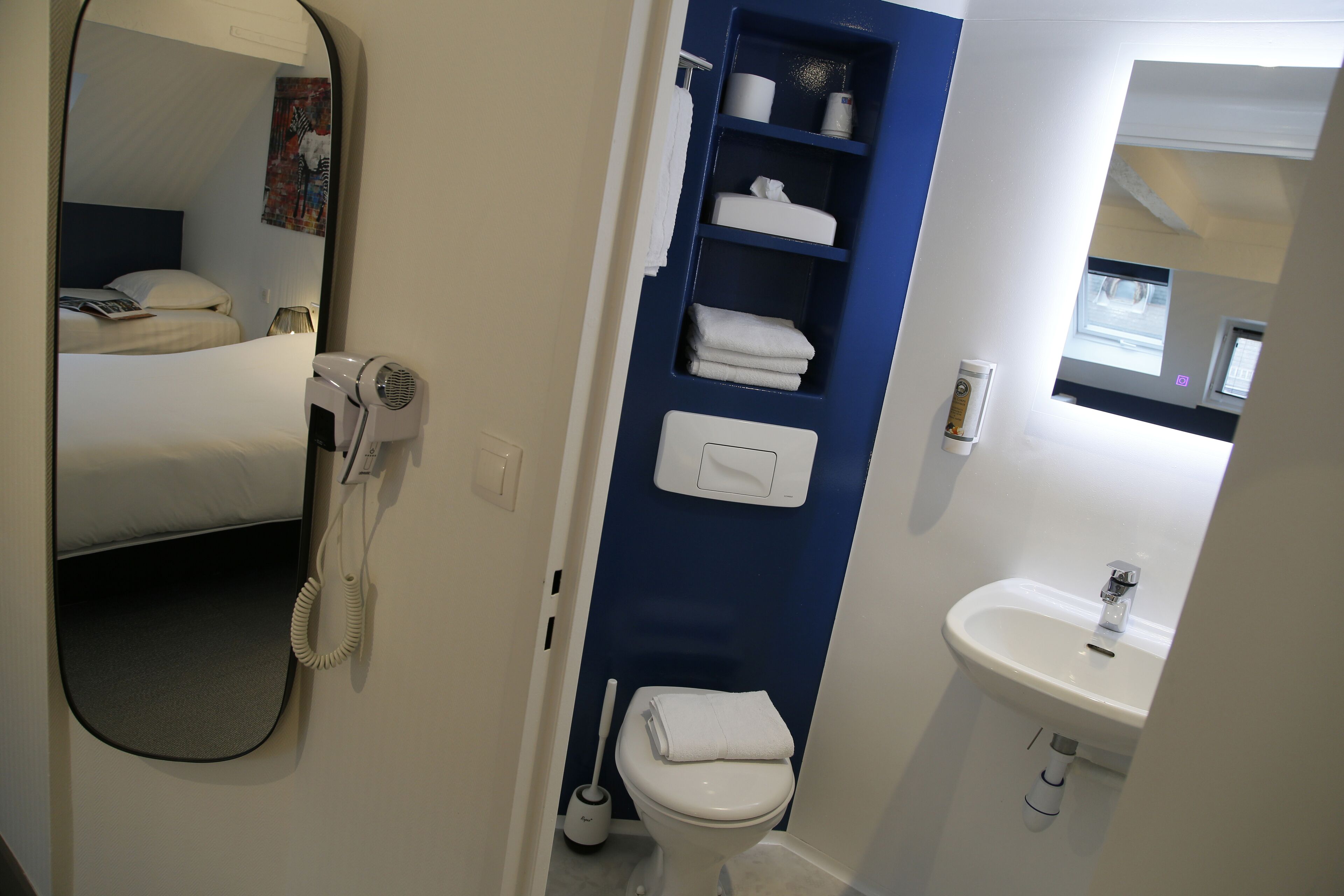 A hotel bathroom with a bed, a hairdryer, a toilet, a sink, and a blue shelf with towels.