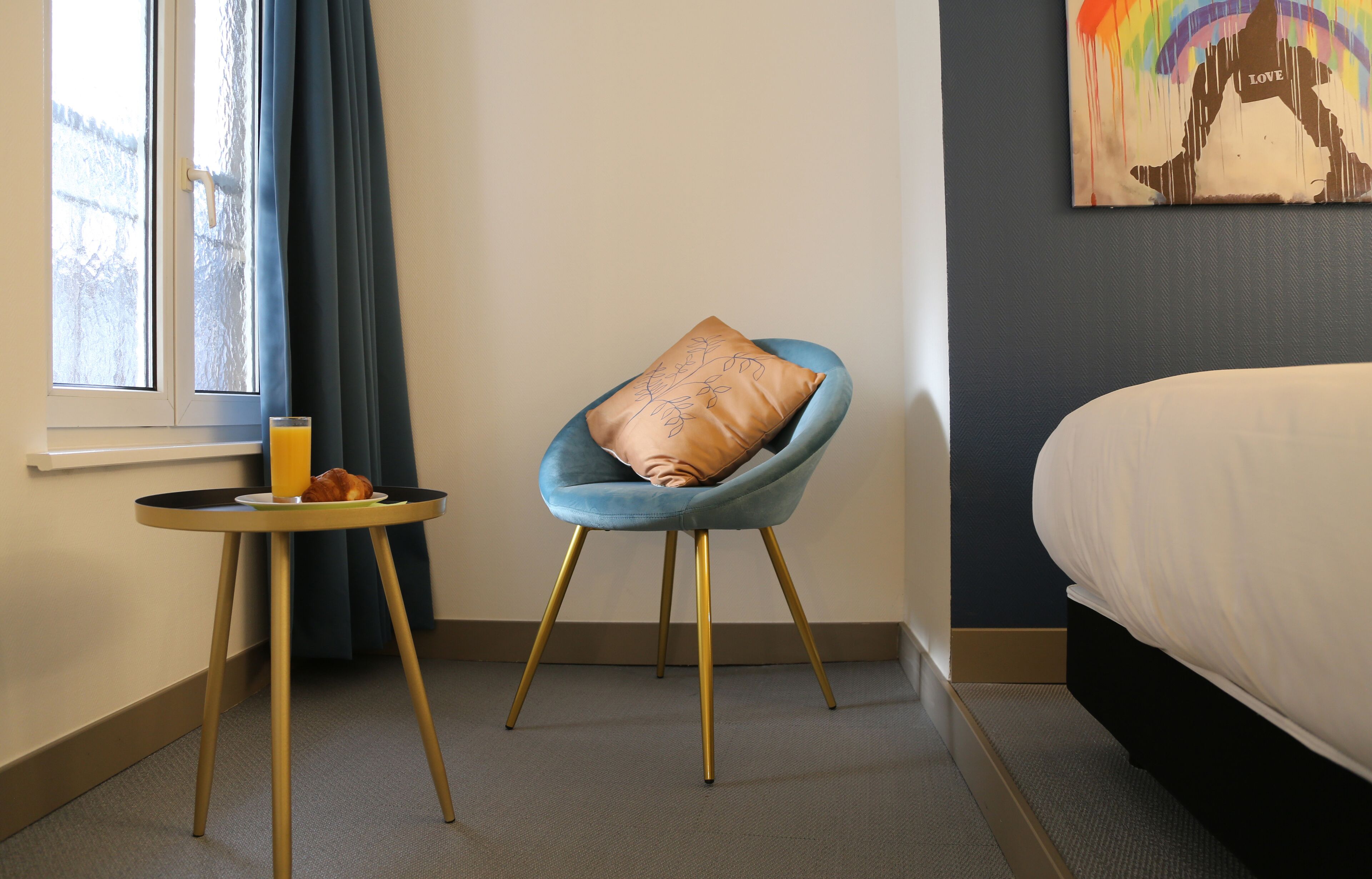 Une chambre d’hôtel avec un lit, un fauteuil bleu avec un coussin marron, une petite table ronde avec un verre de jus d’orange et un croissant, et une fenêtre avec des rideaux bleus.