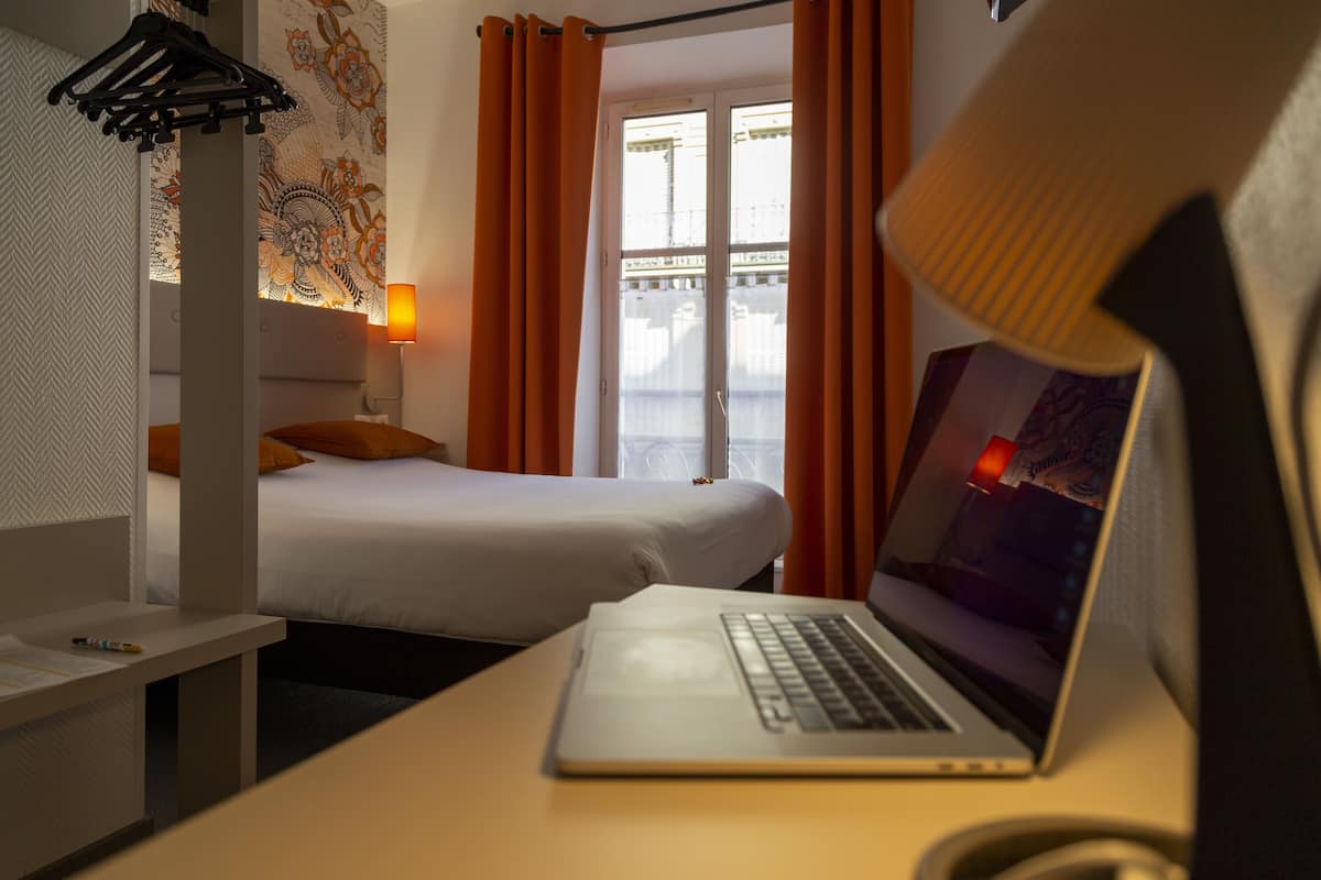 A hotel room with a bed, a desk with a laptop, a lamp, and a window with curtains.
