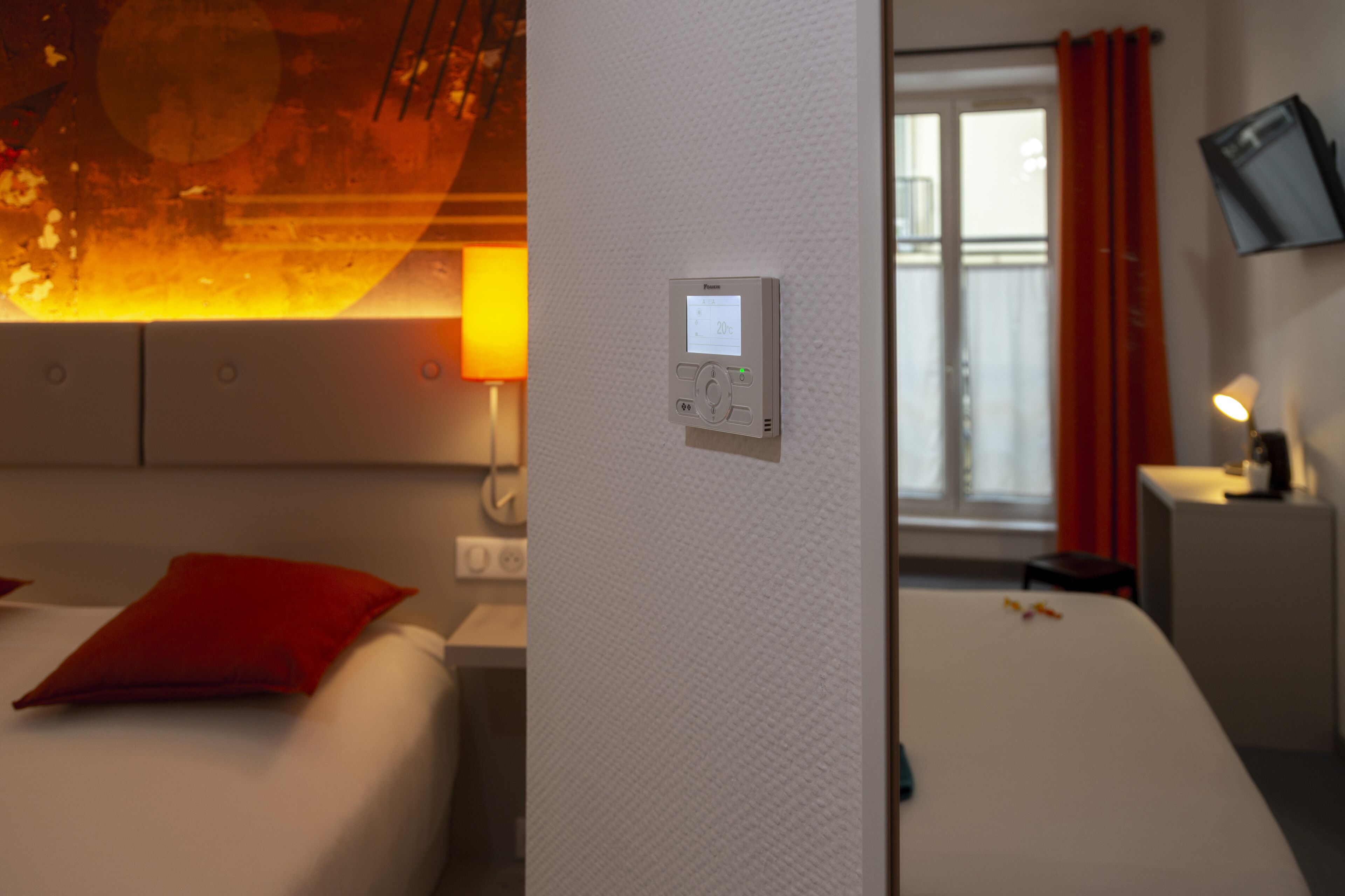 Ein Hotelzimmer mit Bett, Wandthermostat, an der Wand montiertem Fernseher und einem mit Vorhängen versehenen Fenster.