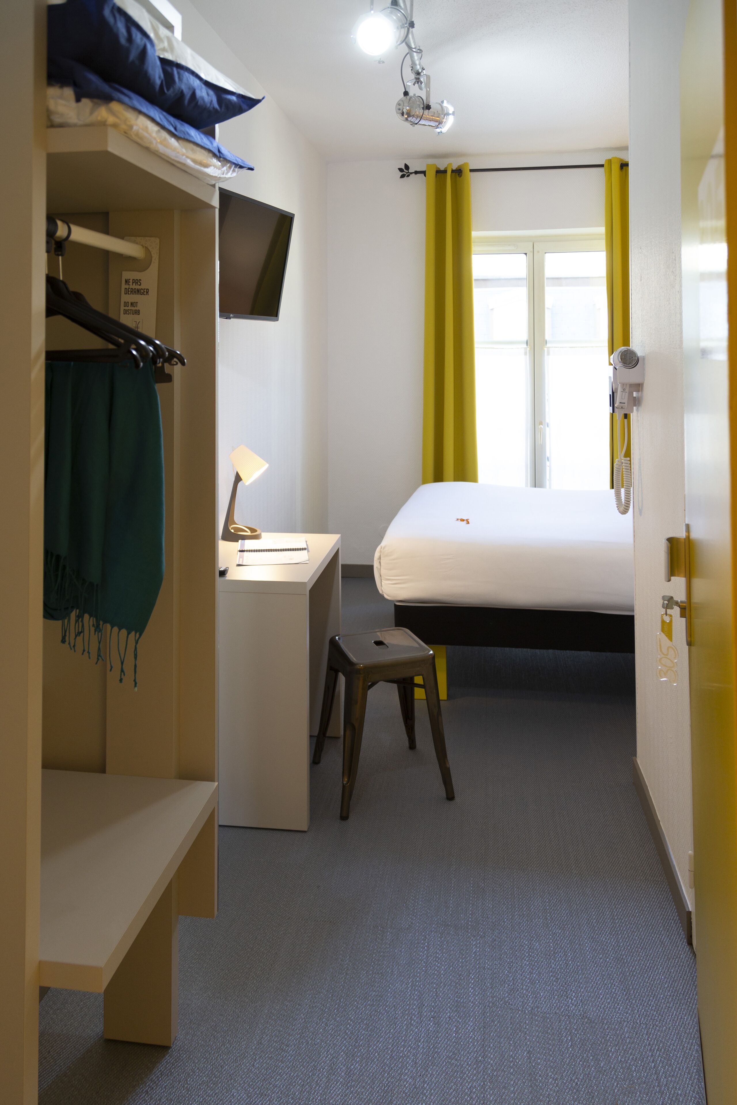 A hotel room with a bed, a desk with a lamp, a chair, a TV mounted on the wall, and a window with yellow curtains.