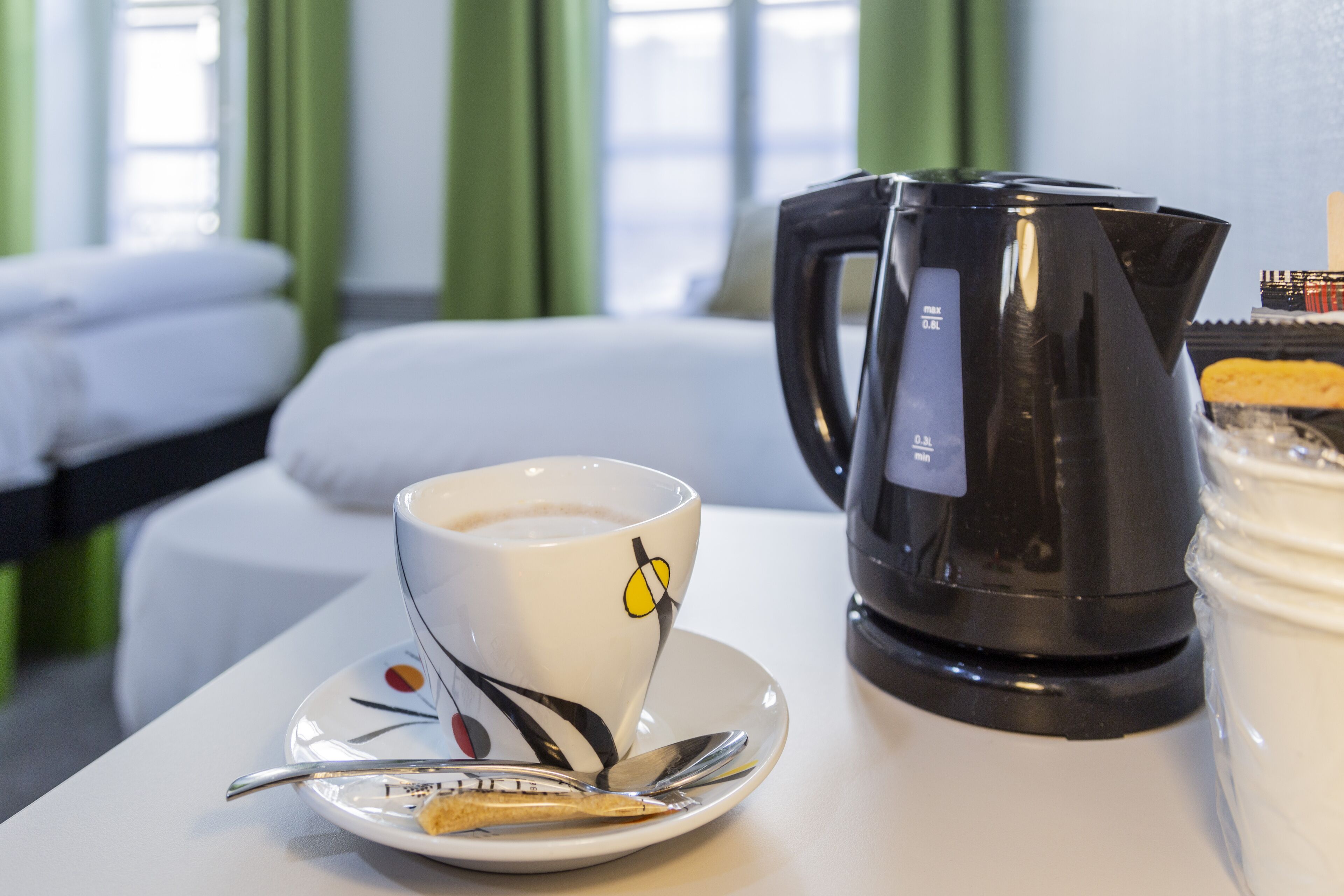 Habitación de hotel con una cama, una taza de café sobre un platillo, una cuchara, una tetera eléctrica negra y un envase de crema.