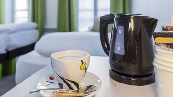 Habitación de hotel con una cama, una taza de café sobre un platillo, una cuchara, una tetera eléctrica negra y un envase de crema.