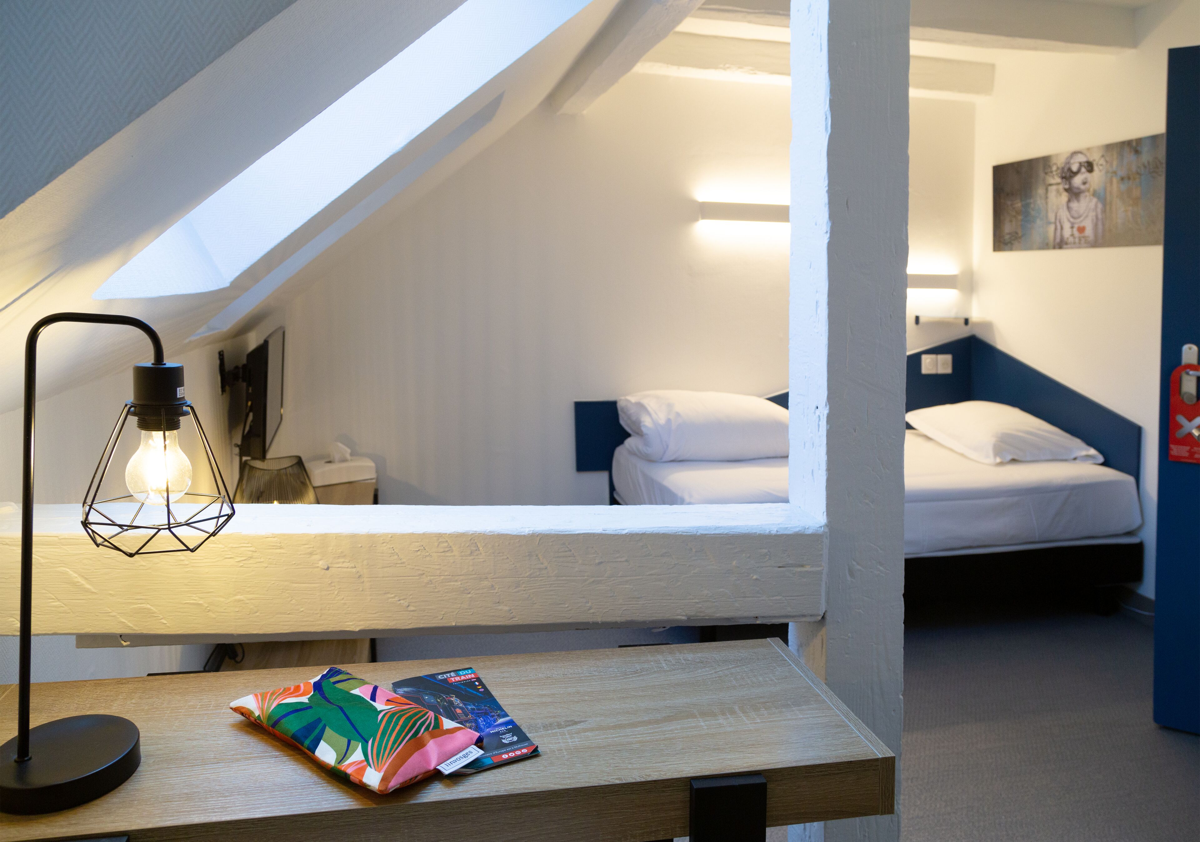 Una habitación pequeña y acogedora con techo inclinado, una cama individual, una mesita de noche con lámpara y un cojín colorido.