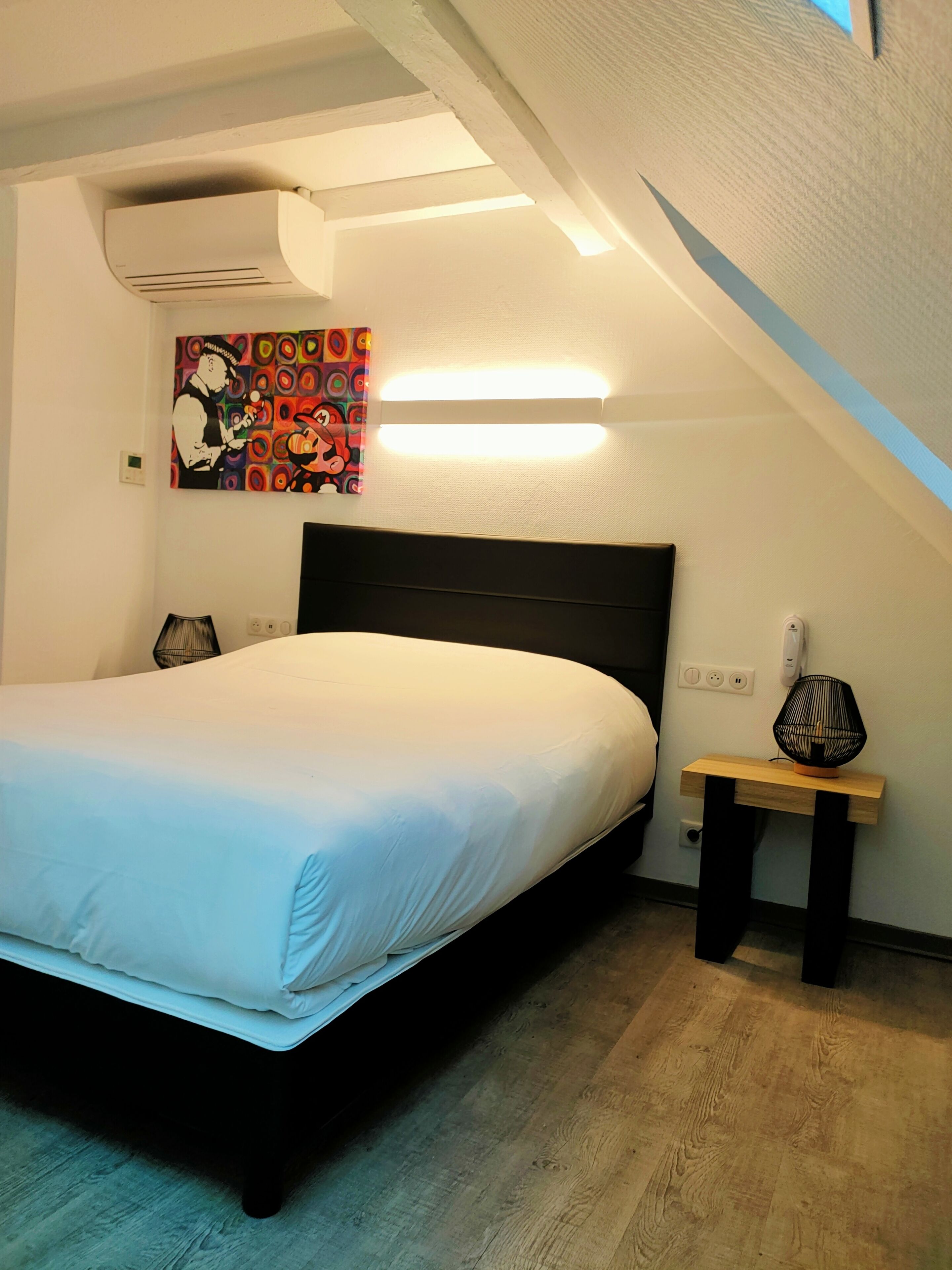 Une chambre à coucher comprenant un lit, une table de chevet, un climatiseur fixé au mur, une œuvre d’art colorée et un luminaire au plafond.