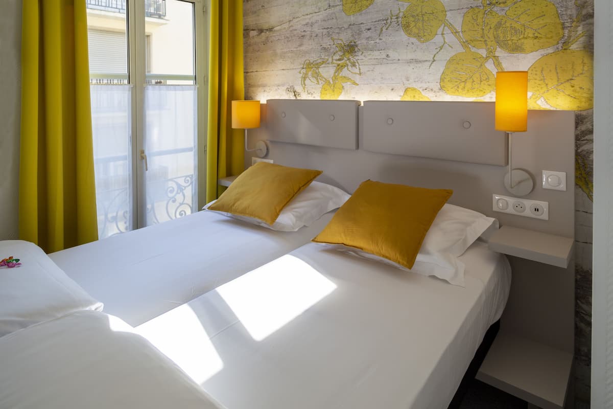 A hotel room with two beds, yellow curtains, a wall with a leaf pattern, and a nightstand with a lamp.