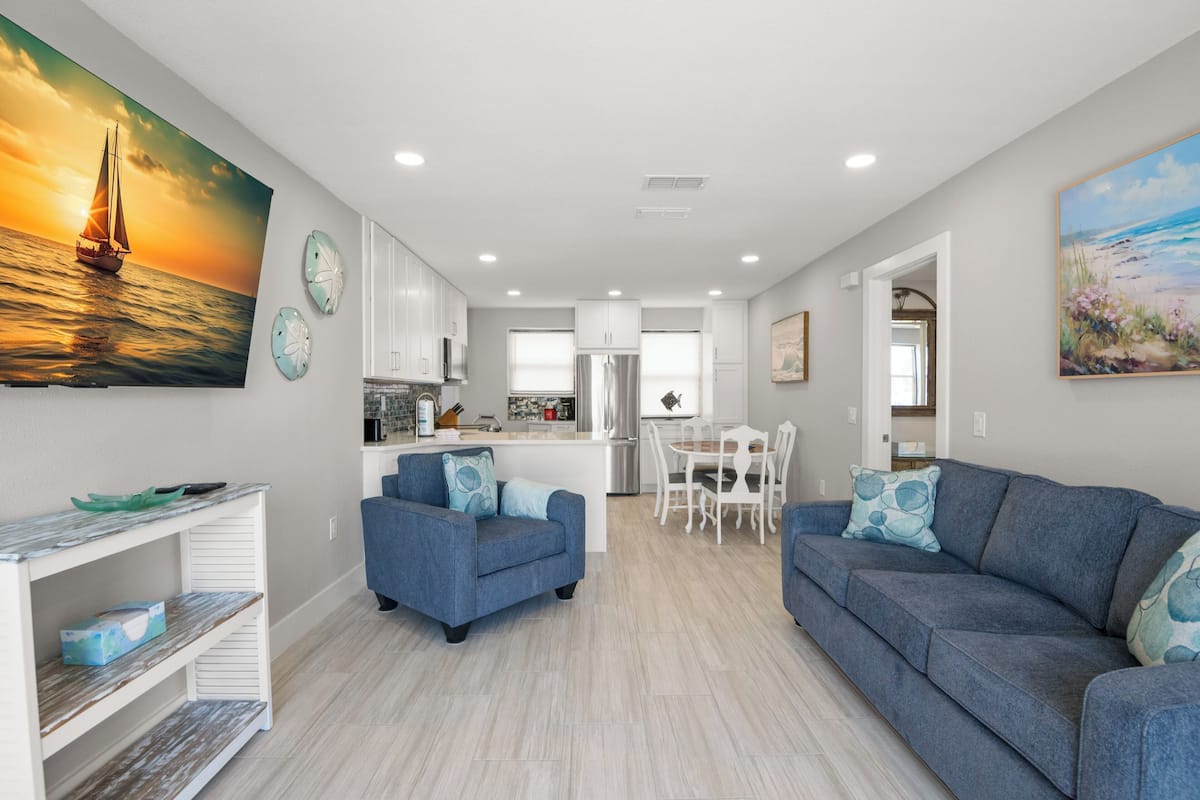 A modern kitchen with white cabinets, a dining area with a white table and chairs, and a living room with a blue sofa and matching cushions.