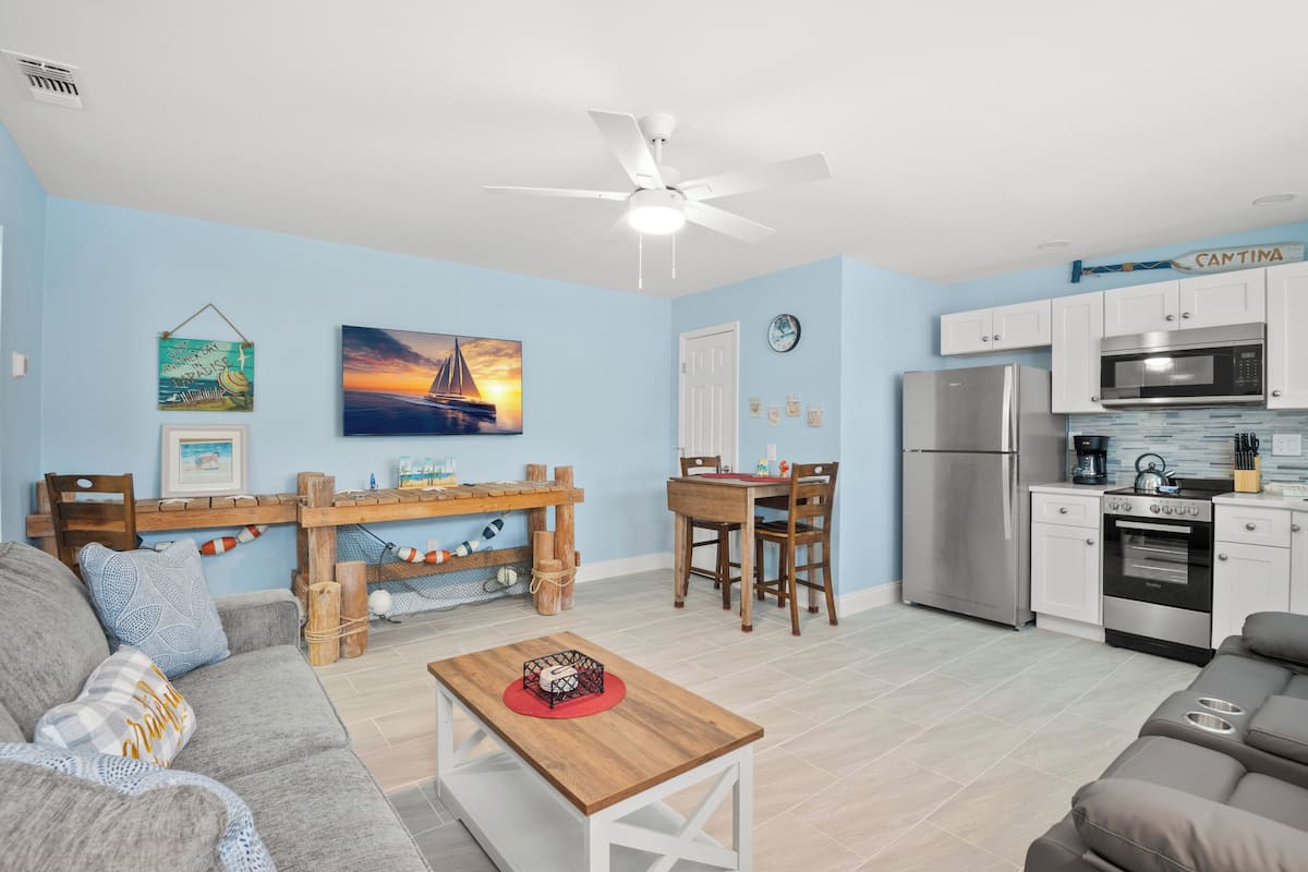 A living room with a sofa, a coffee table, and a dining area with a table and chairs. There is a kitchen with a refrigerator, microwave, and stove. A ceiling fan is visible, and there are wall decorations including a painting and a sign.